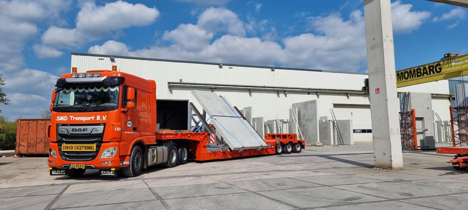 Oranje SMD Transport B.V. DAF vrachtwagen met lage trailer geladen met grote betonnen platen, geparkeerd bij een industriële opslaglocatie.