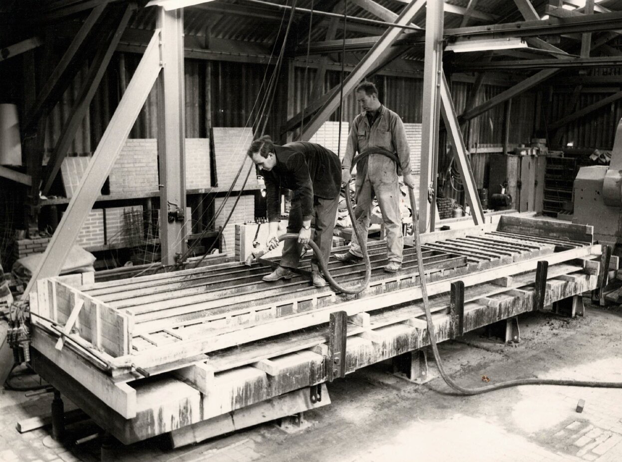 Historische foto circa jaren '70: twee arbeiders werken aan een grote betonvorm in een industriële loods.