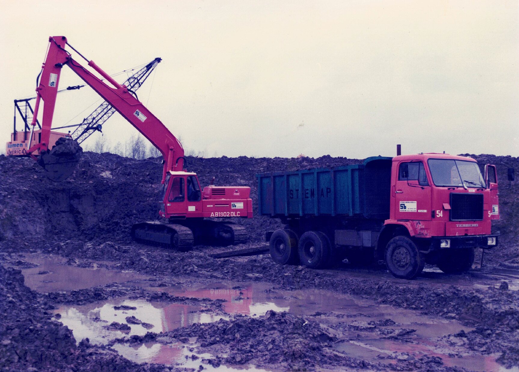 Historische foto circa jaren '80: Stenap graafmachine laadt grond in een kiepwagen op een modderige bouwplaats.