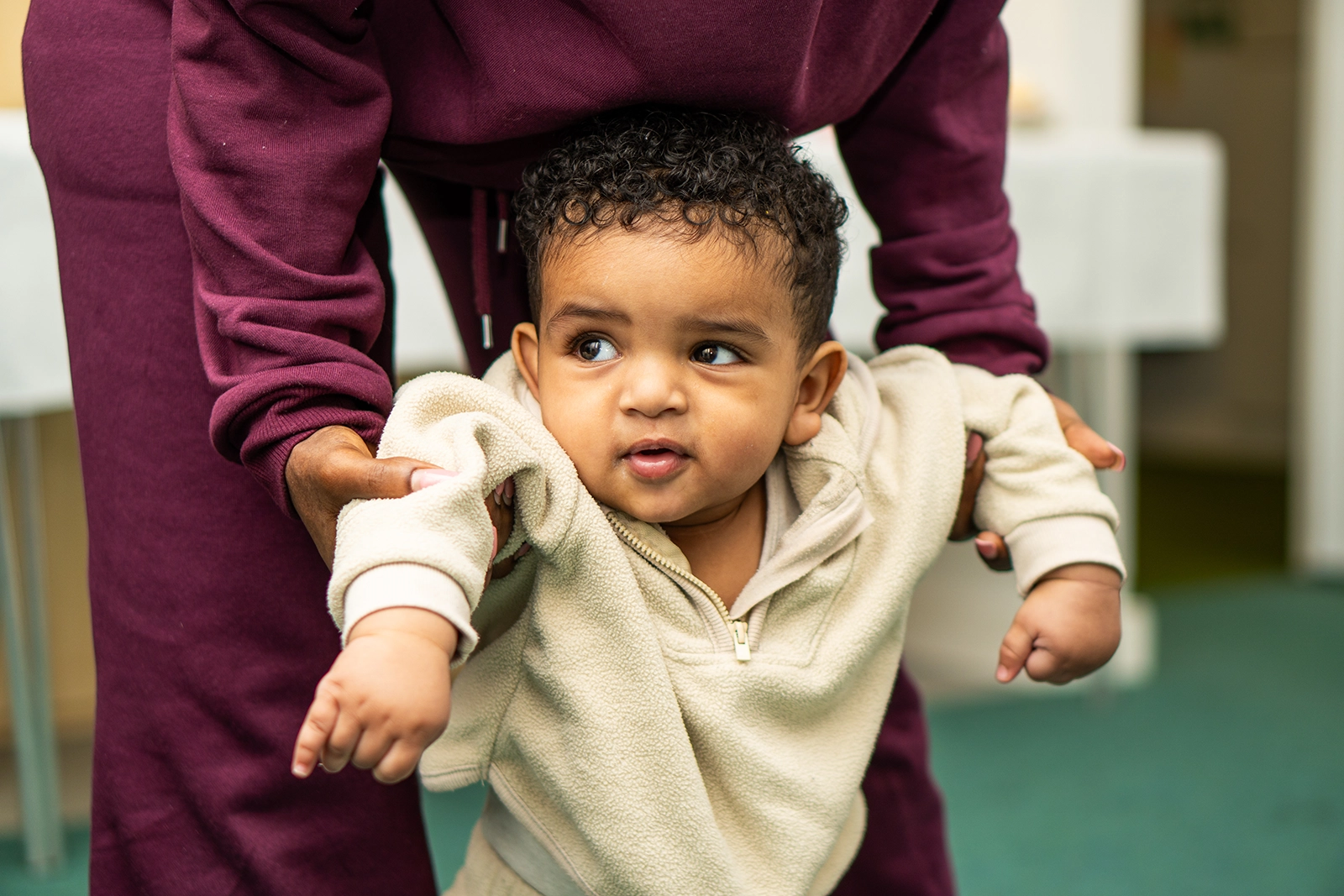 Adult in maroon outfit supporting a baby with curly hair wearing a beige fleece sweater, helping them stand or walk indoors.