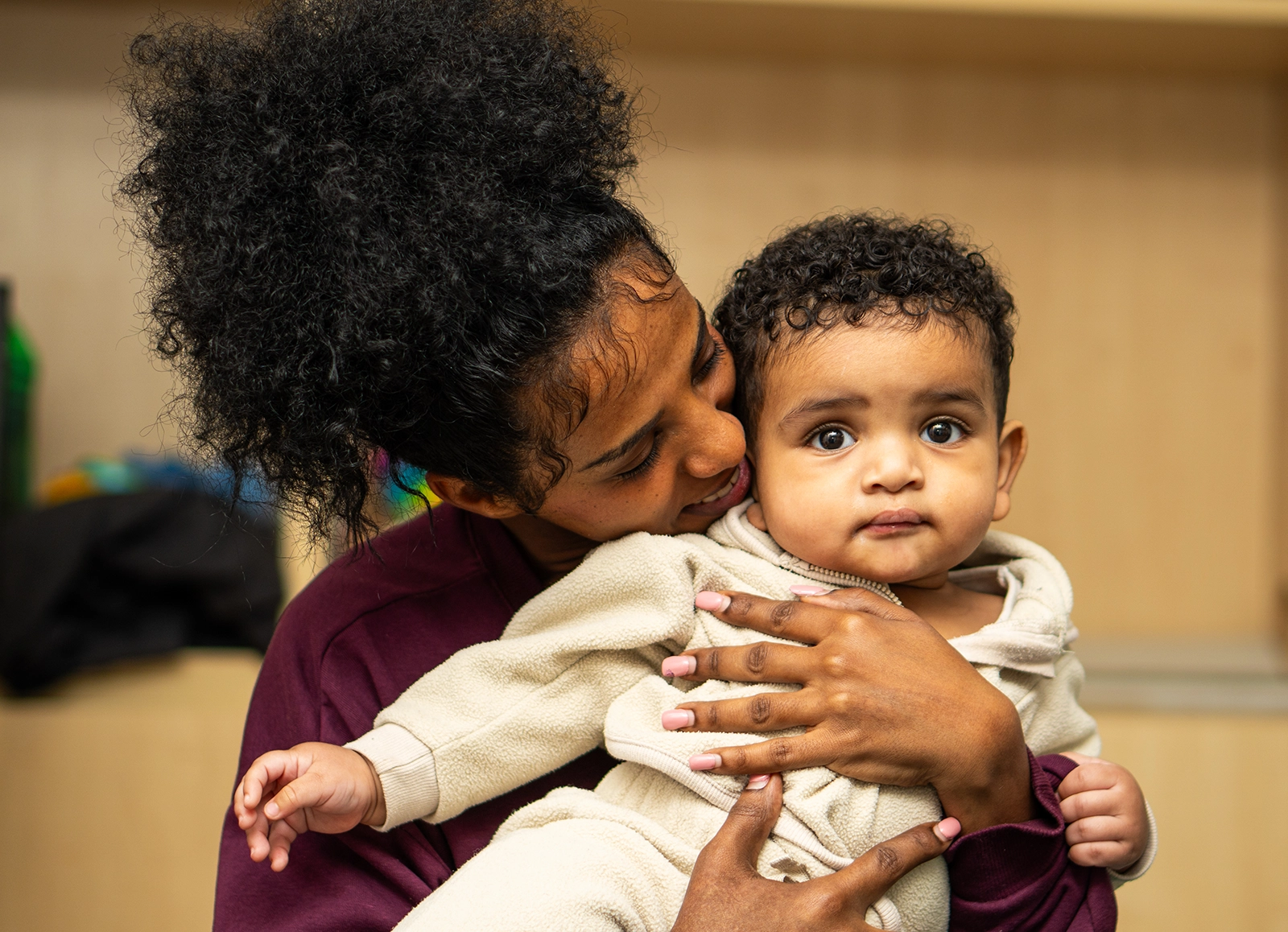 Woman with curly hair holding and embracing a baby wearing a beige outfit.