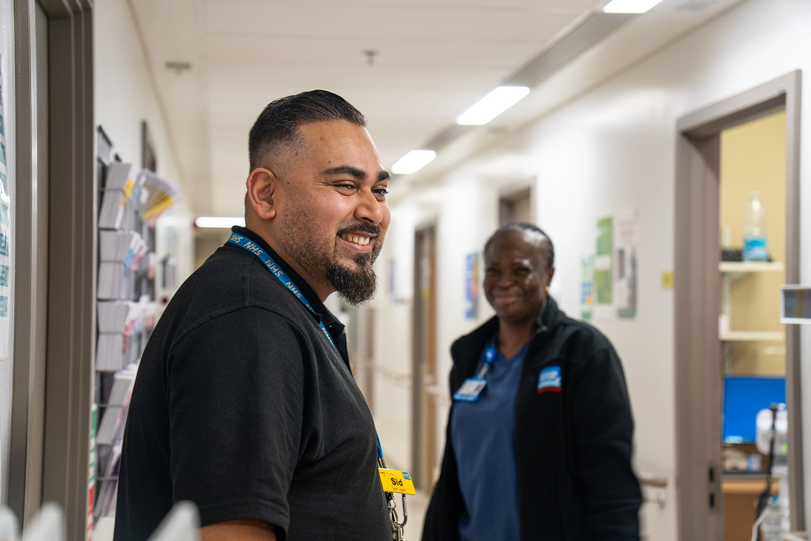 Two healthcare workers smiling in a hospital corridor, one wearing an NHS lanyard and name badge.