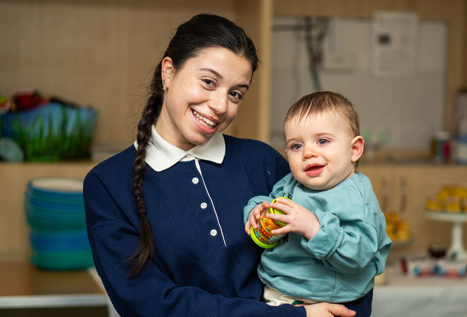 Smiling woman with braided hair holding a baby who is playing with a colorful toy inside a room.