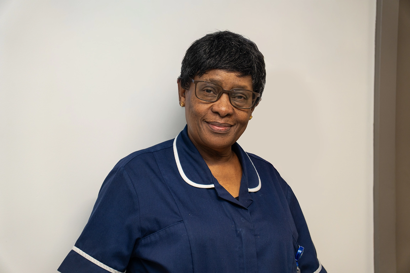 Smiling female nurse wearing glasses and a navy blue uniform with white trim, standing against a plain background.