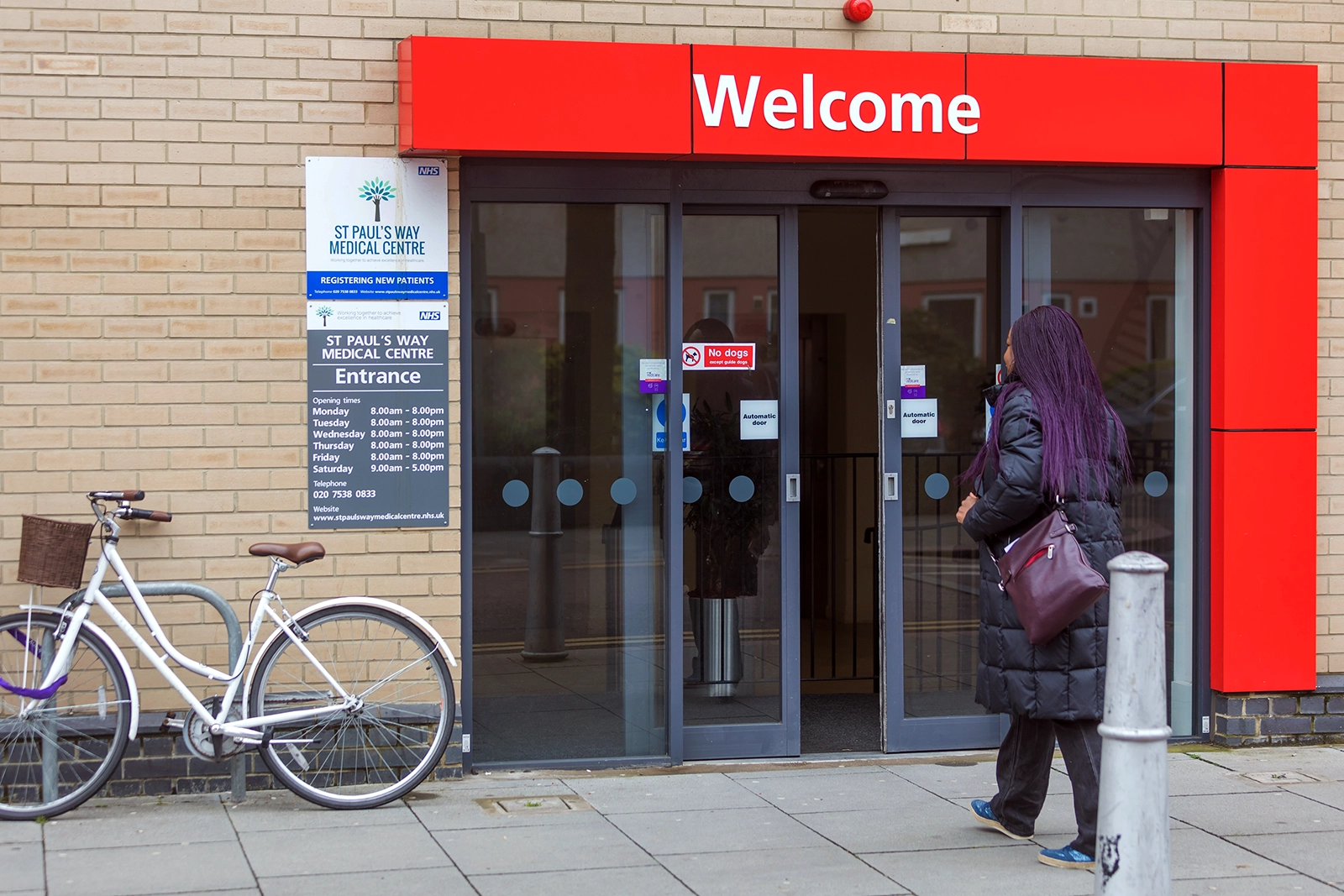 Woman with long purple hair entering St Paul's Way Medical Centre through automatic glass doors under a red welcome sign.