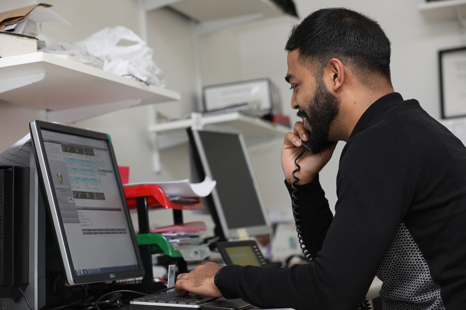 Man with a beard talking on a phone while typing on a keyboard at a desk with dual monitors and organized paper trays.