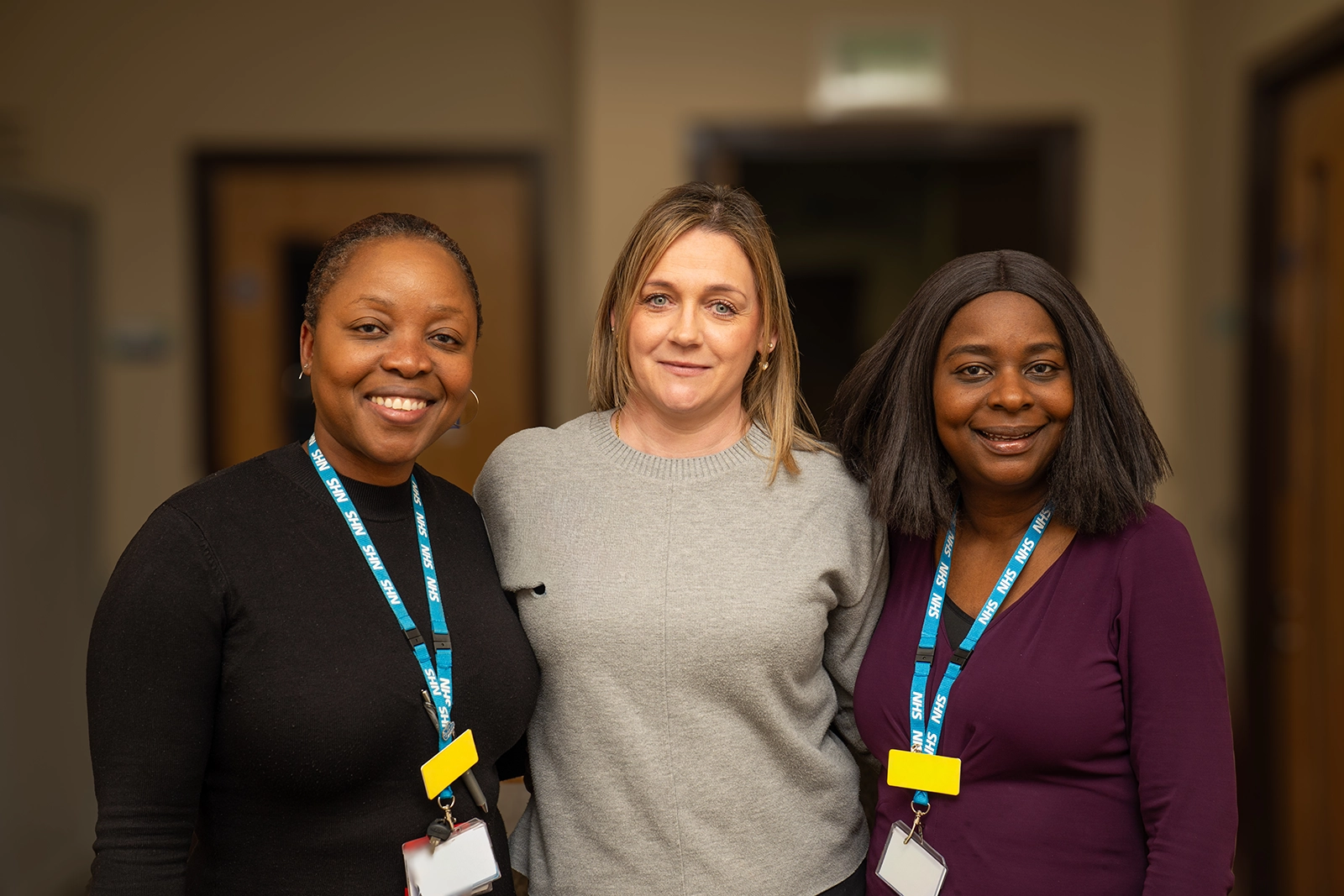 Three smiling women standing indoors, two wearing NHS lanyards and ID badges.