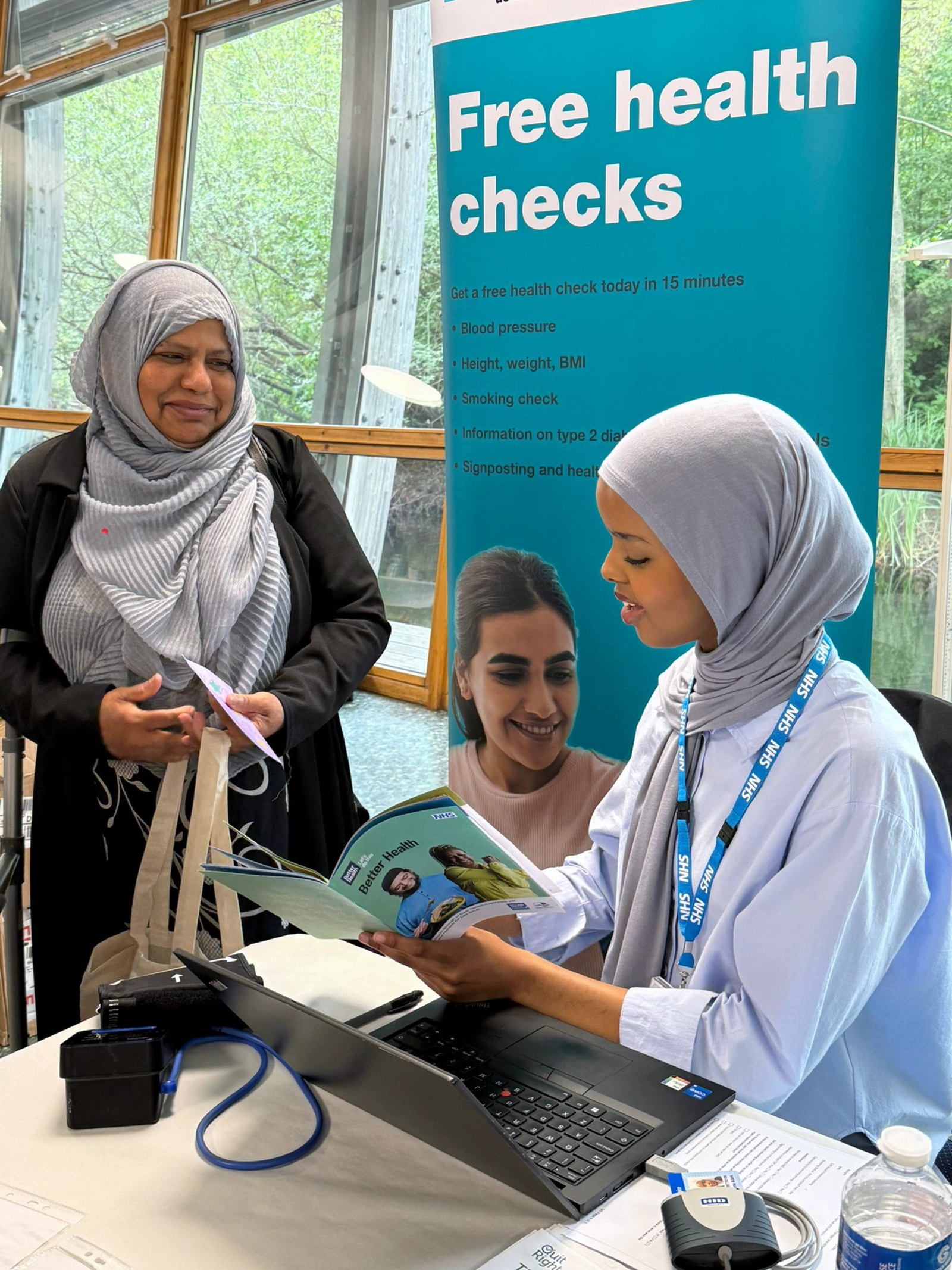 Woman in a grey hijab receiving health information from a healthcare worker in a grey hijab at a free health checks booth with a laptop and pamphlets.