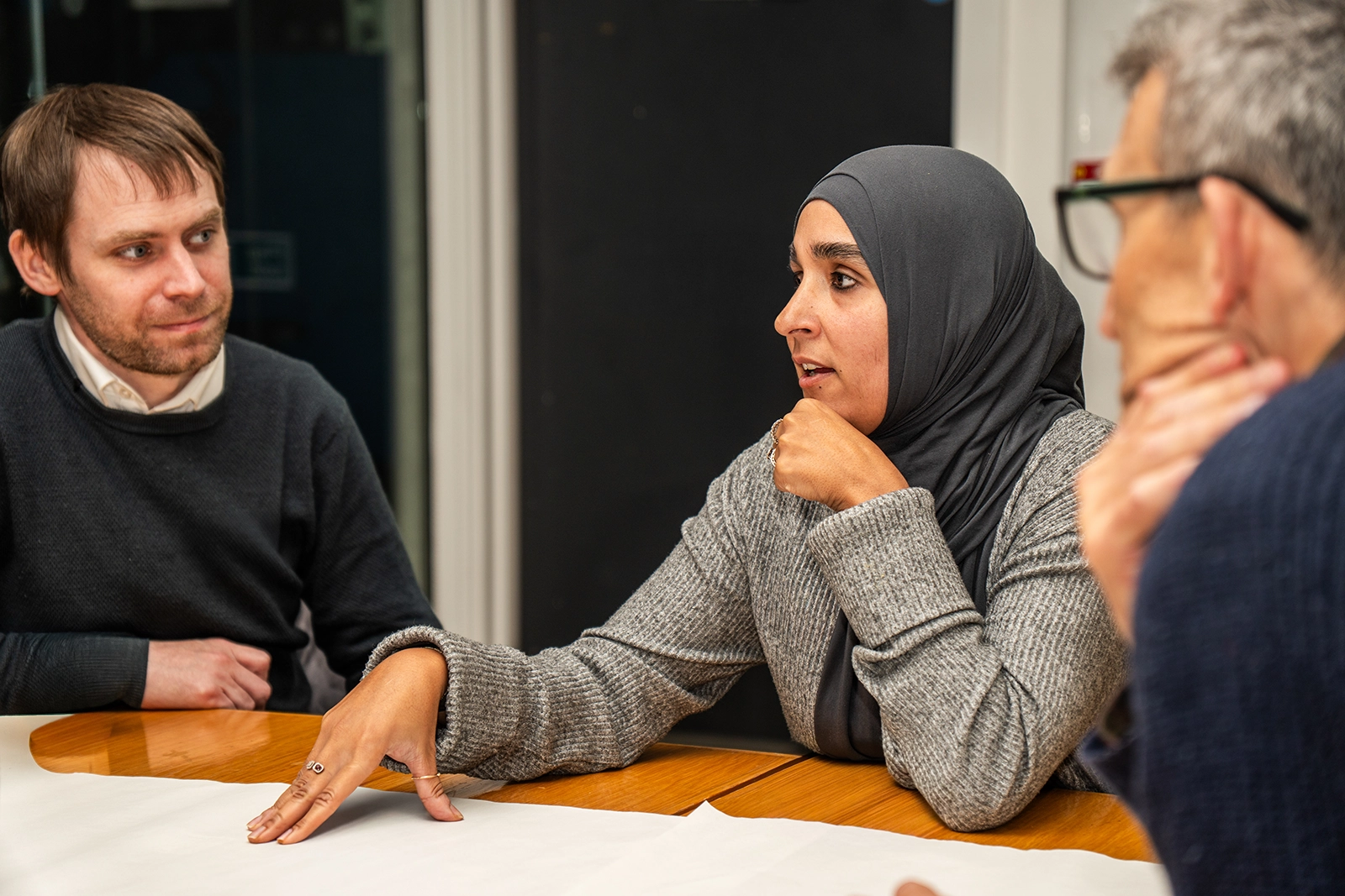 A woman wearing a hijab and gray sweater talking at a table with two men during a discussion.