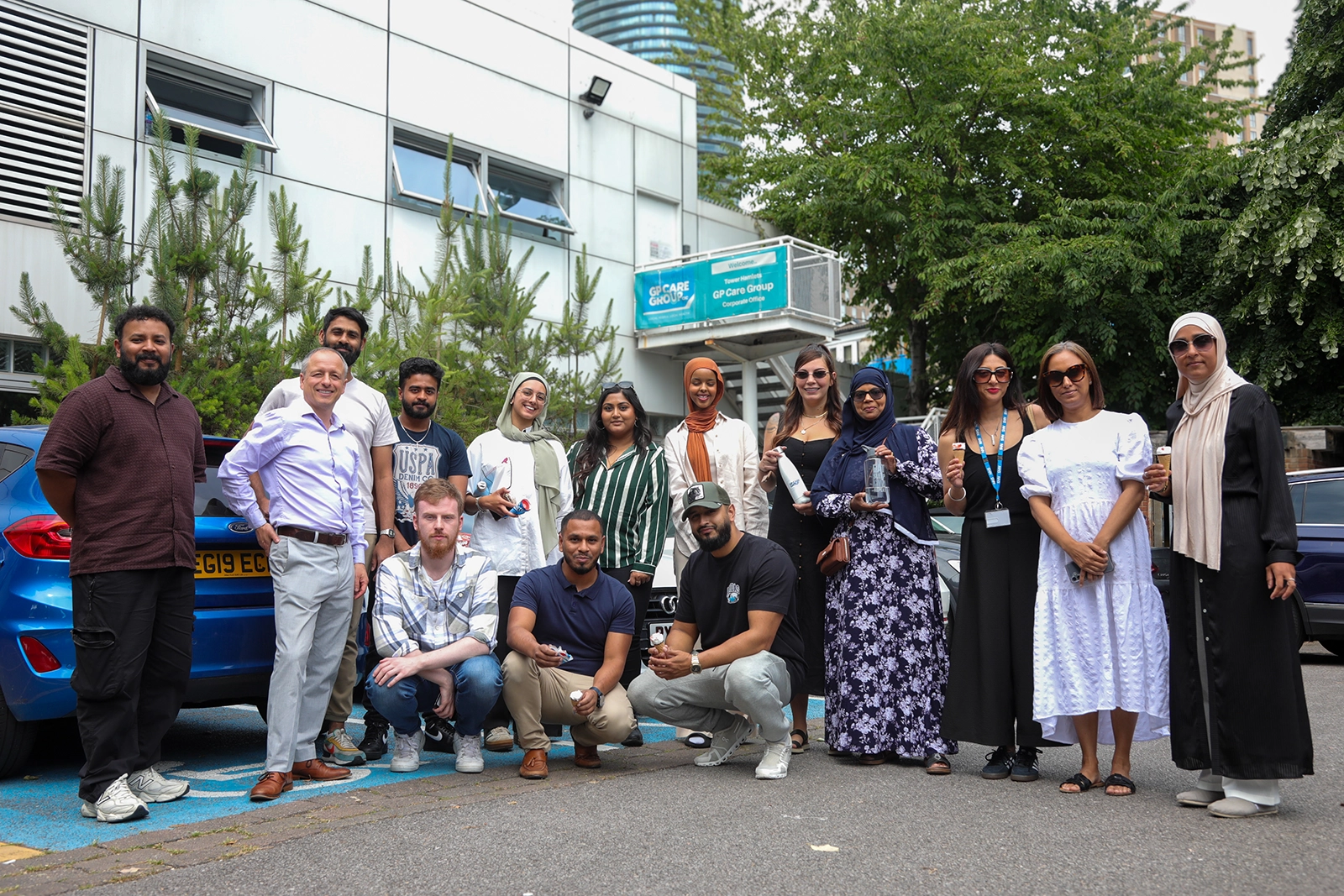A diverse group of people standing and squatting outside a building with a GP Care Group sign, holding ice cream cones and bottles, in a casual group photo.