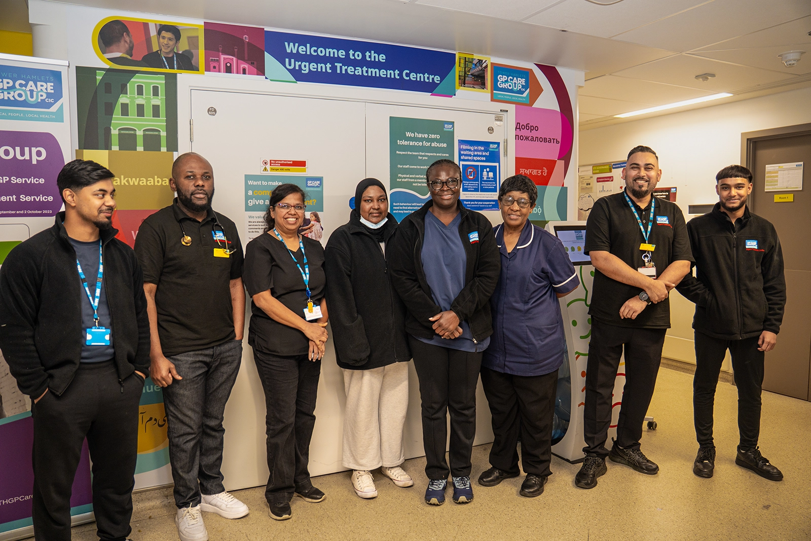 Group of eight diverse healthcare workers standing and smiling inside the Urgent Treatment Centre with welcoming signs in the background.