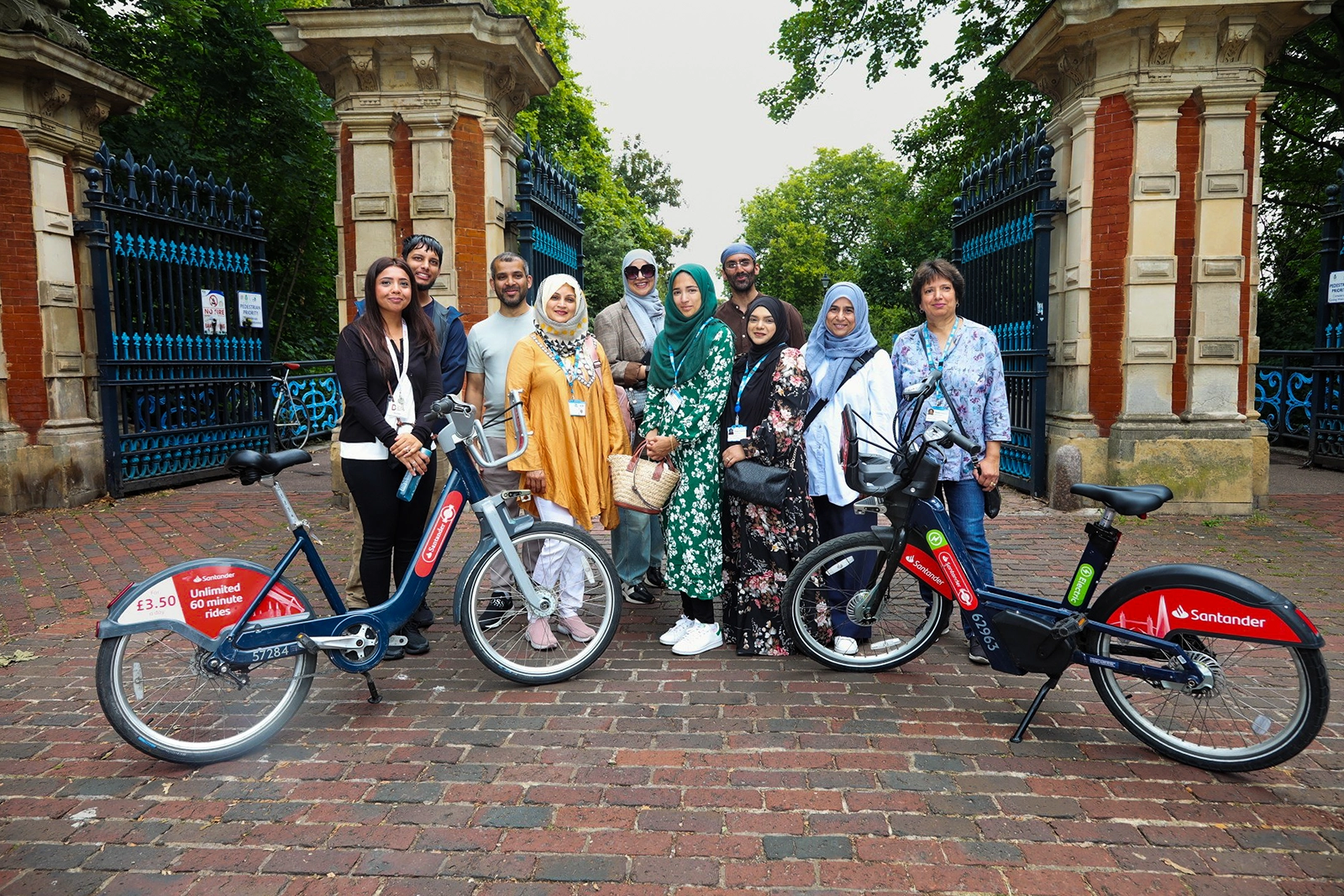 Group of ten people standing behind two Santander rental bikes on a brick pathway with ornate iron gates and greenery in the background.