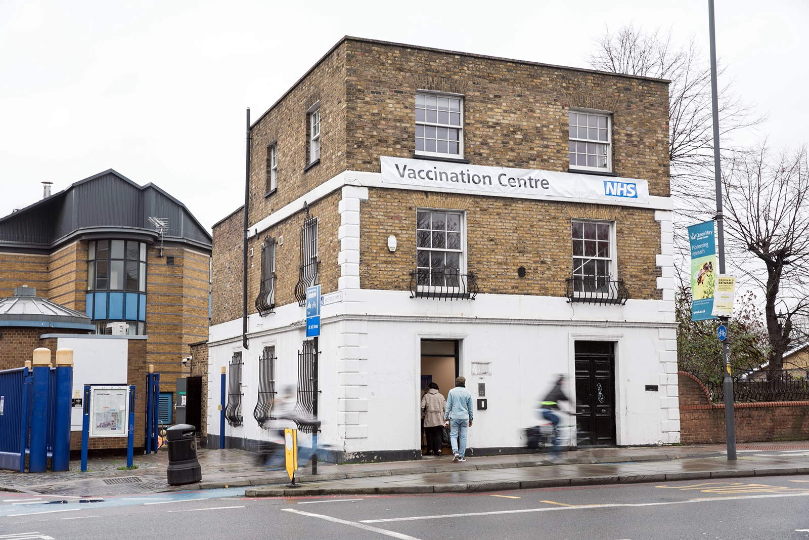 Brick building with a white lower facade labeled Vaccination Centre with NHS logo, people entering, and cyclists passing by.