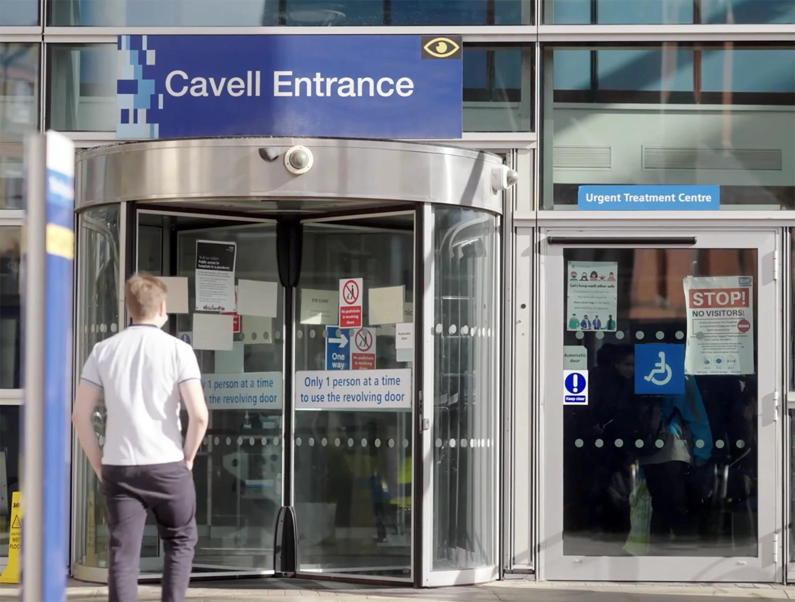 Person walking towards revolving glass door labeled 'Cavell Entrance' with adjacent door marked 'Urgent Treatment Centre' and signs about entry restrictions.