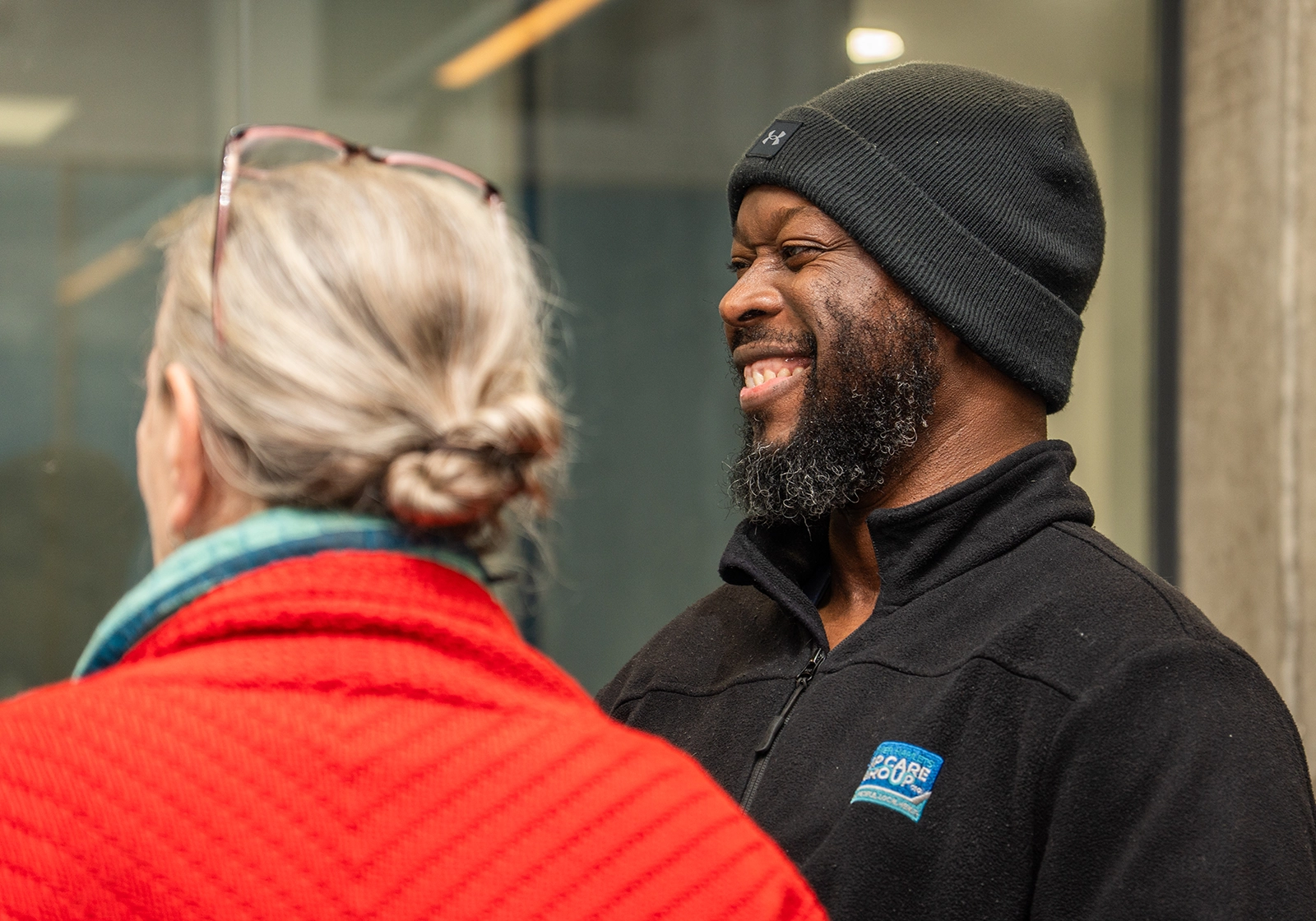 Smiling man in black beanie and fleece jacket talking to a woman wearing a red sweater and teal scarf.