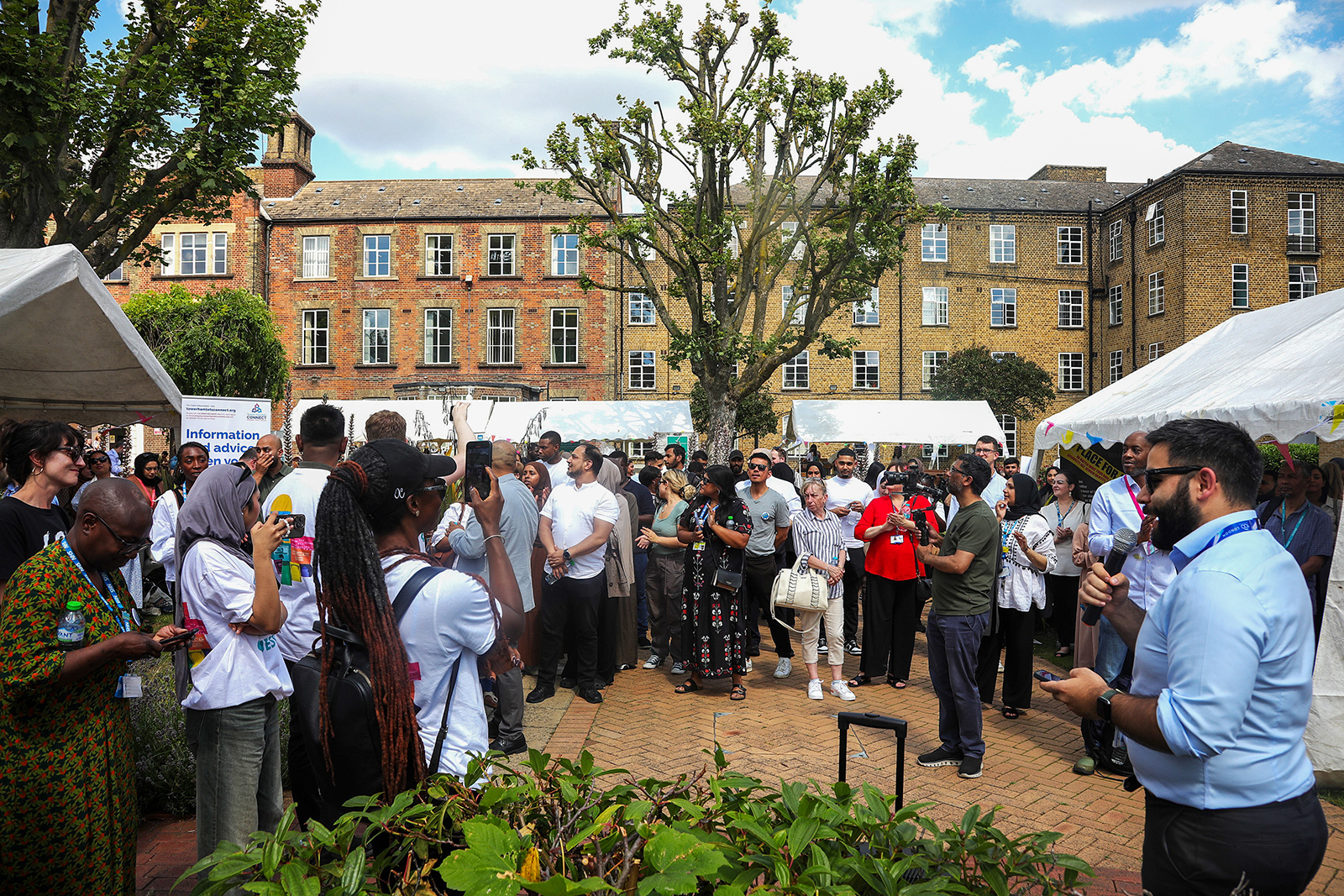 A diverse group of people gathered outdoors at an event with tents, listening to a man speaking into a microphone.