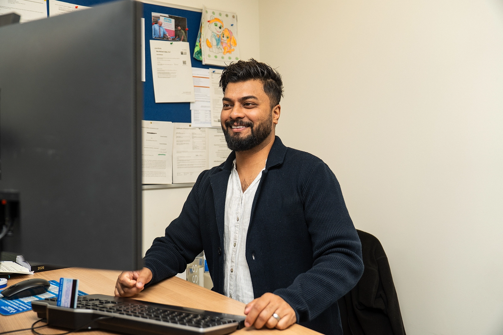 Smiling man with beard sitting at desk using desktop computer in office.