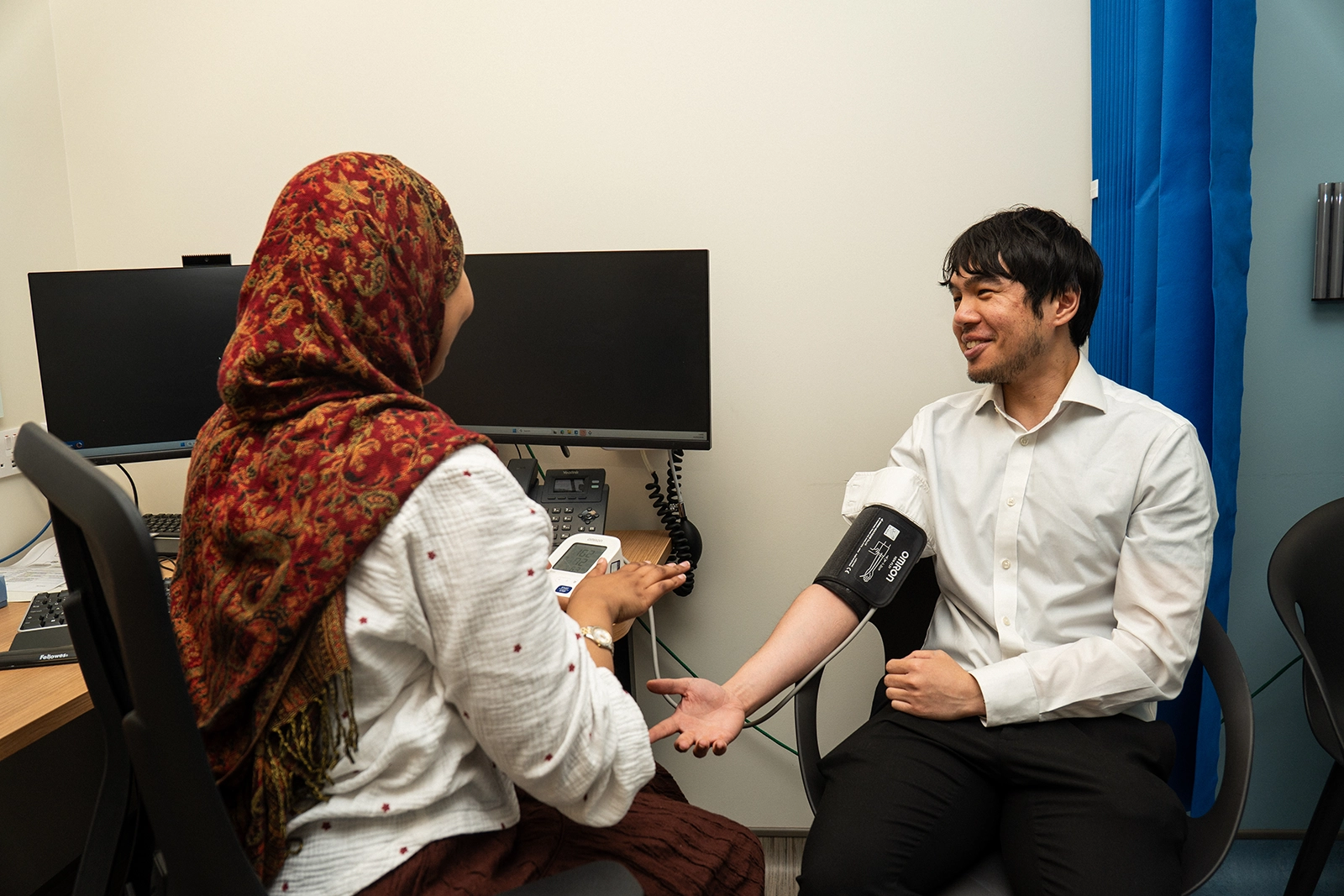 Healthcare professional in a red patterned headscarf measuring the blood pressure of a smiling man wearing a white shirt and black pants in a clinical office.