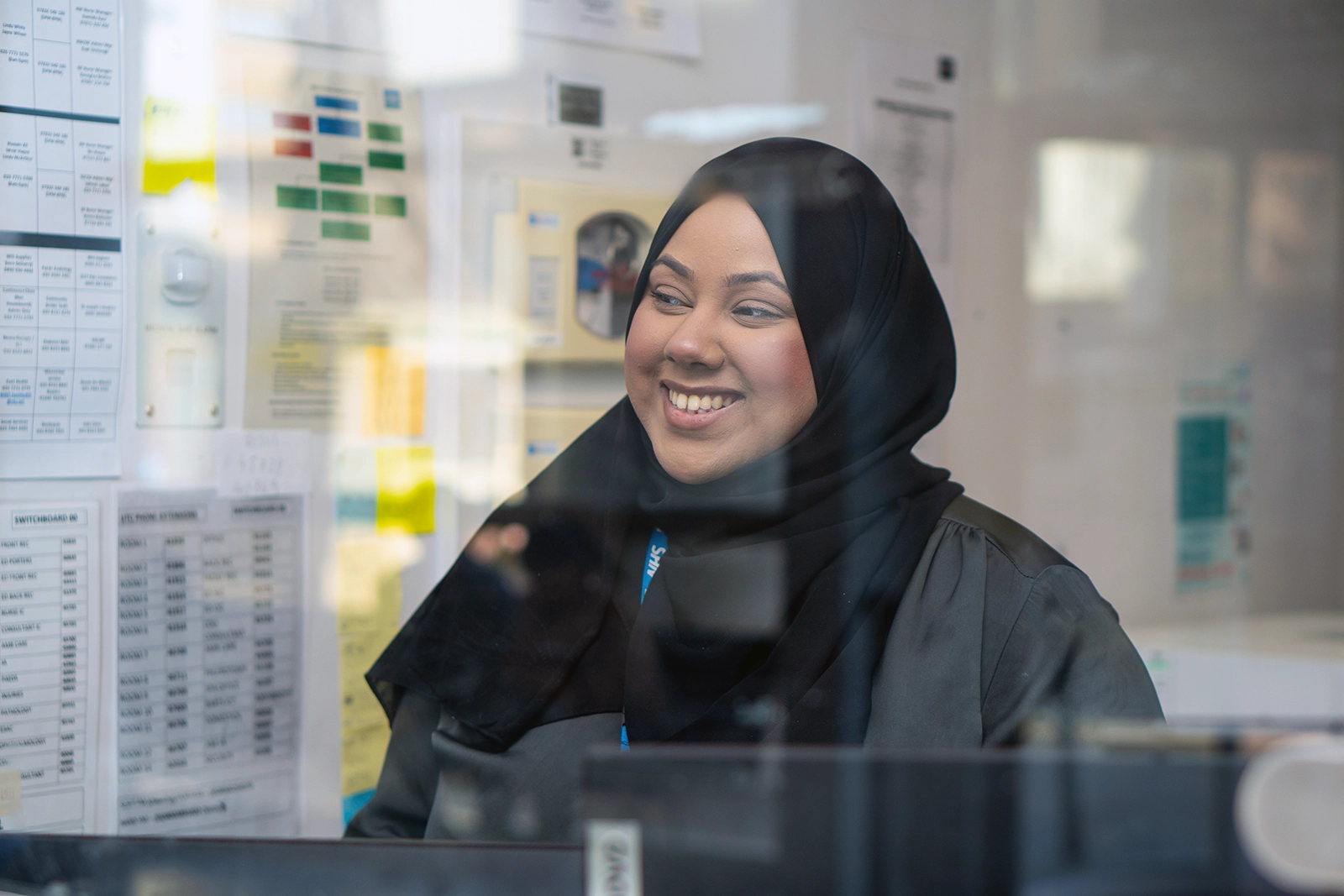 Smiling woman wearing a black hijab, seen through a glass window with office documents in the background.
