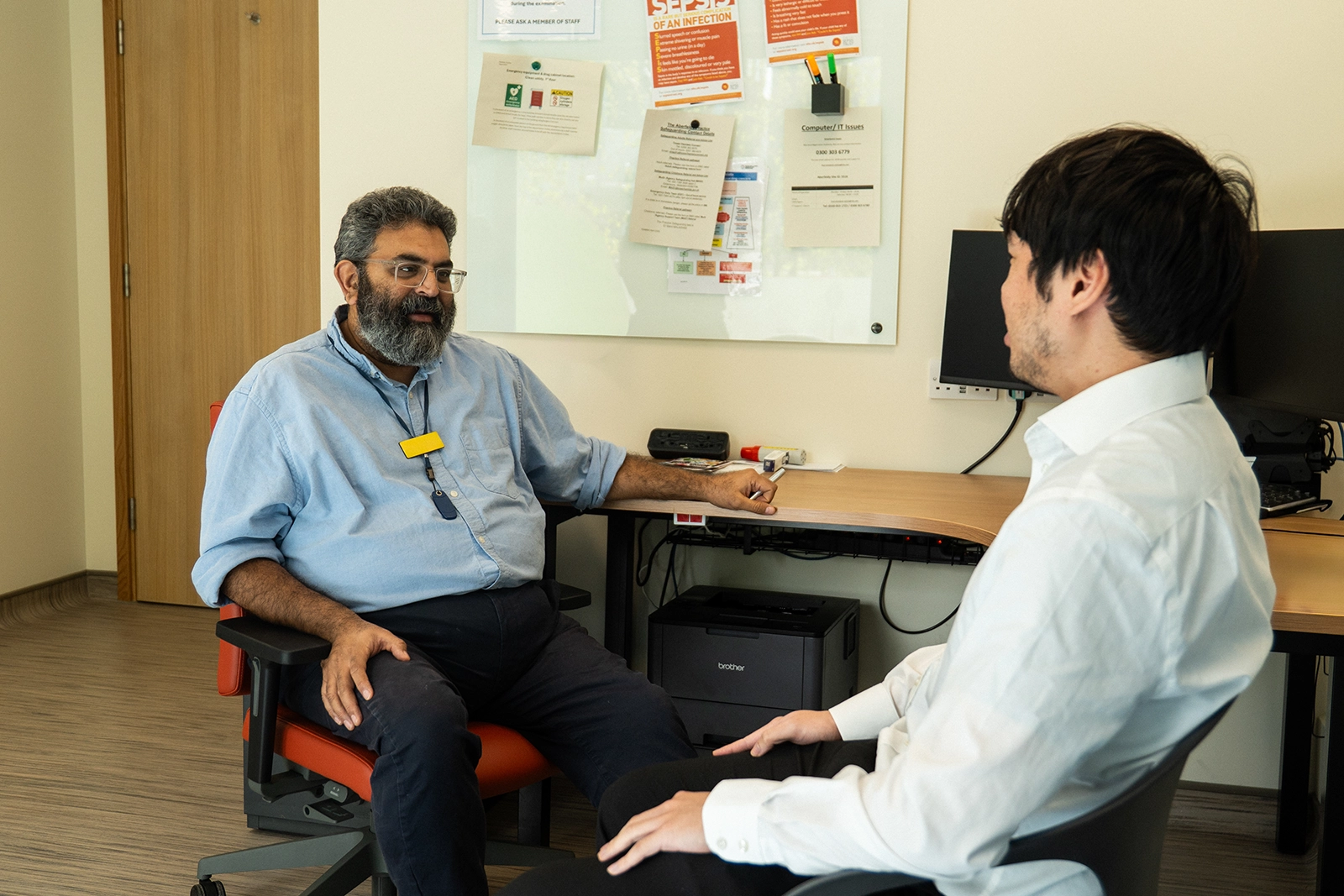 Two men seated and having a conversation in an office with a desk, computer monitors, and a whiteboard with papers.