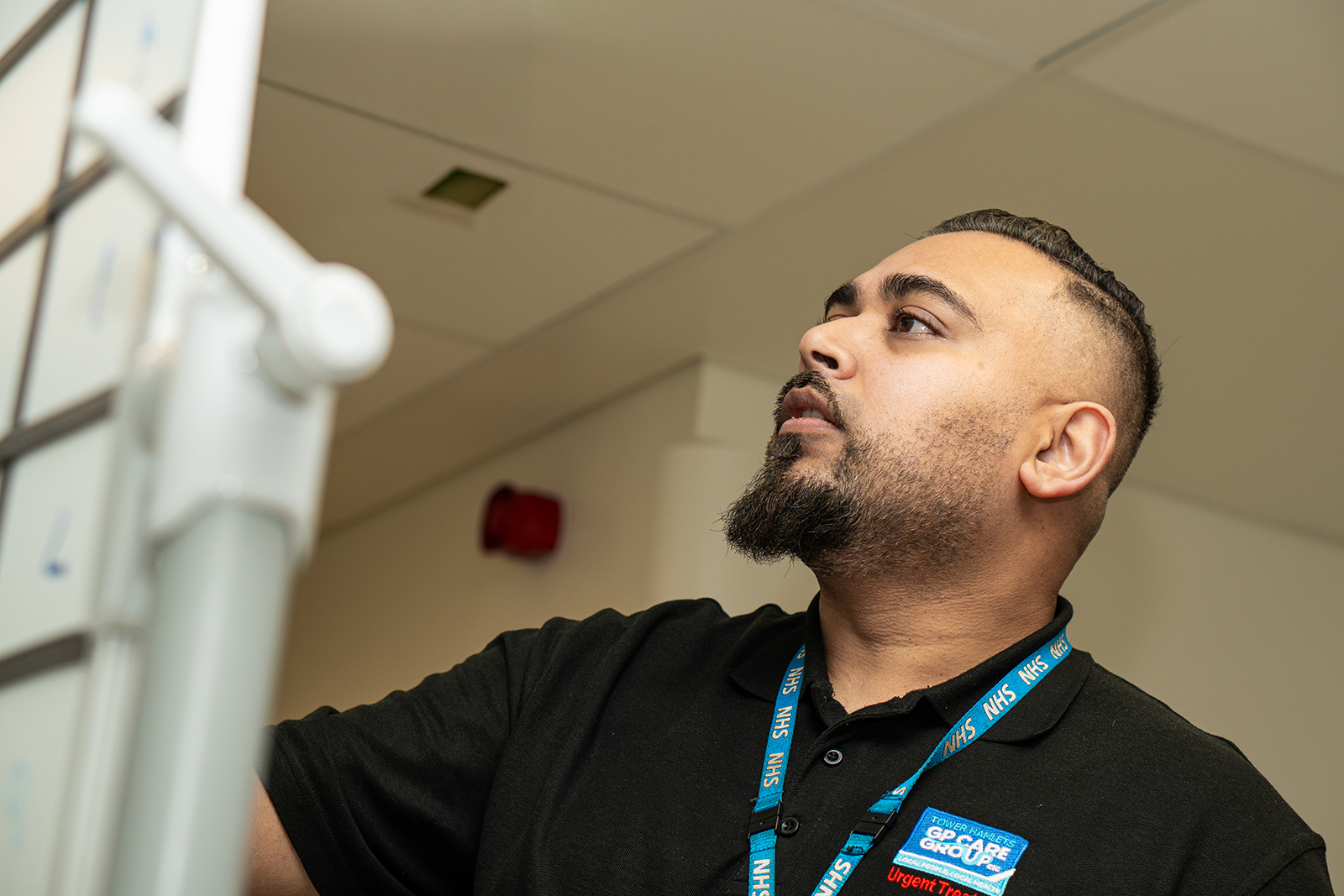 Bearded man wearing a black polo and NHS lanyard looking upwards indoors.