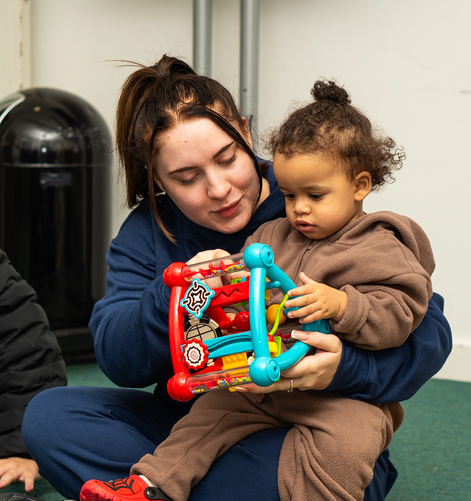 Woman in navy blue hoodie sitting on the floor holding a toddler dressed in brown playing with a colorful activity toy.
