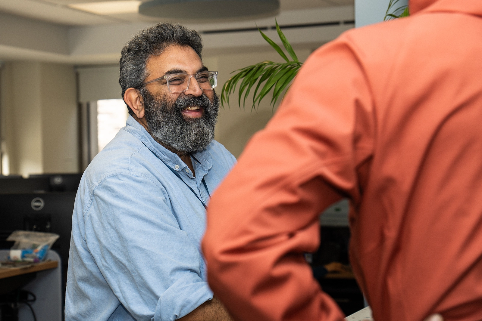 Smiling man with glasses and a beard wearing a light blue shirt, engaging in conversation with another person in an orange shirt.