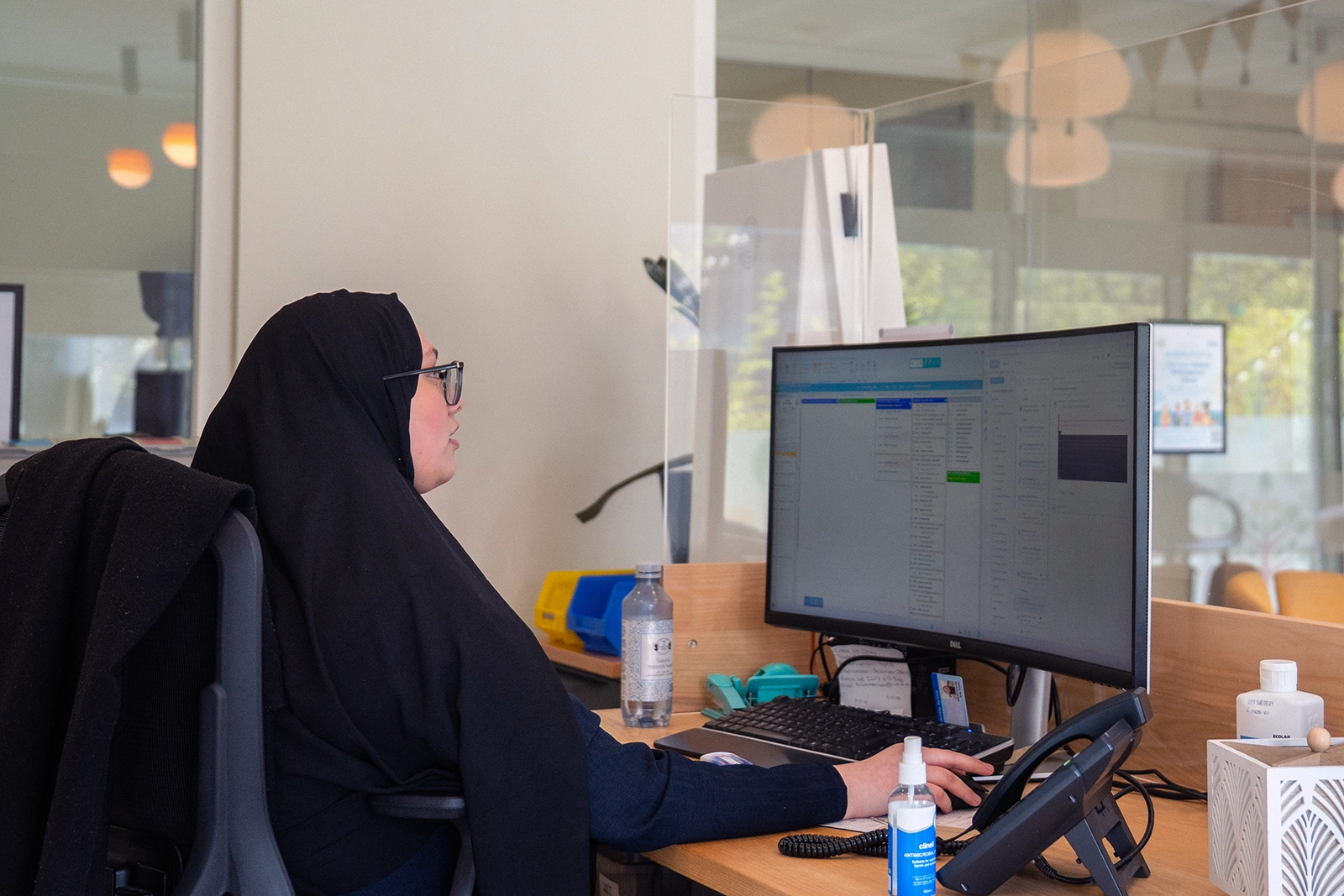 Woman wearing a black headscarf and glasses working on a computer in an office with a transparent partition.