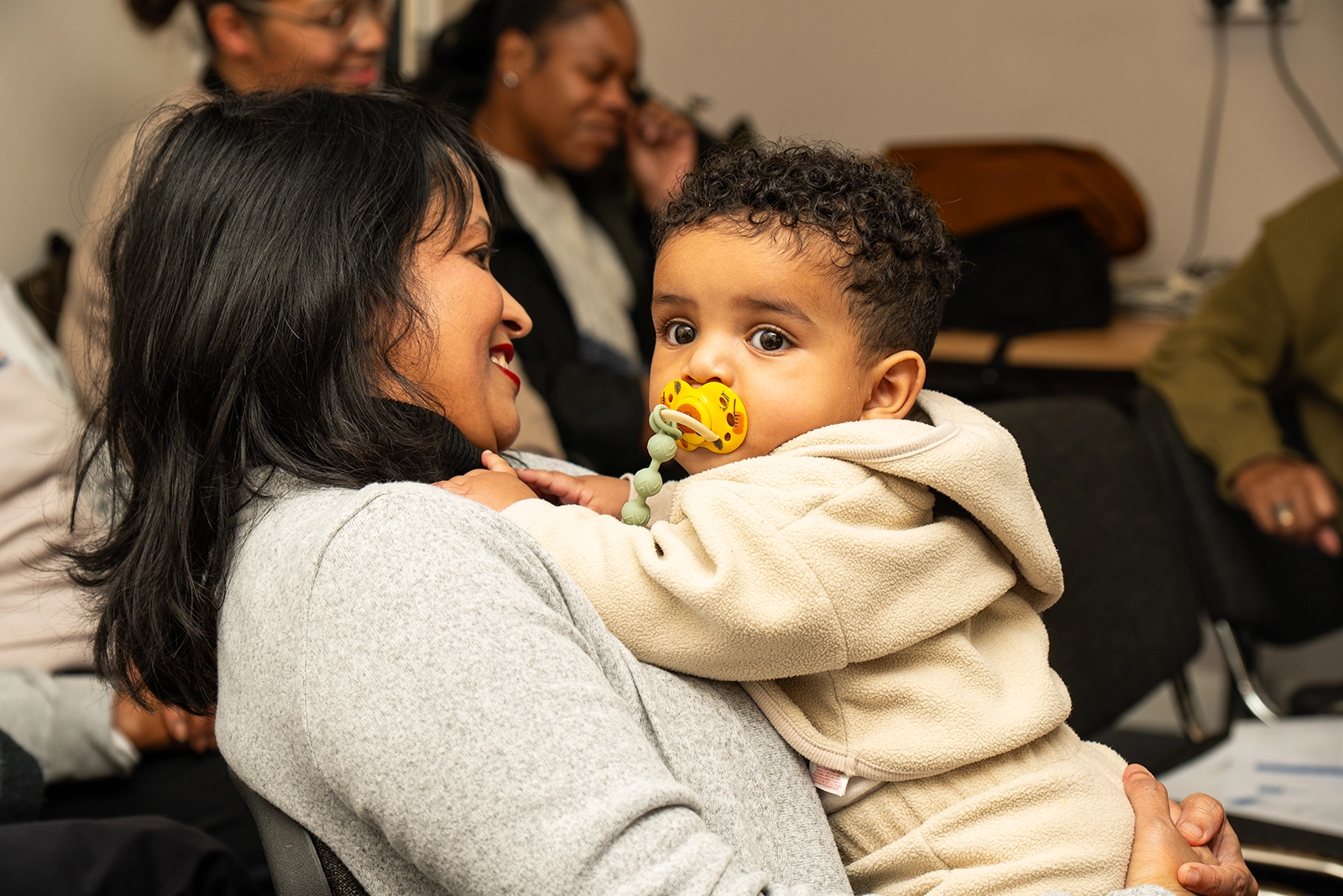 Woman smiling while holding a baby with a pacifier indoors among other seated people.