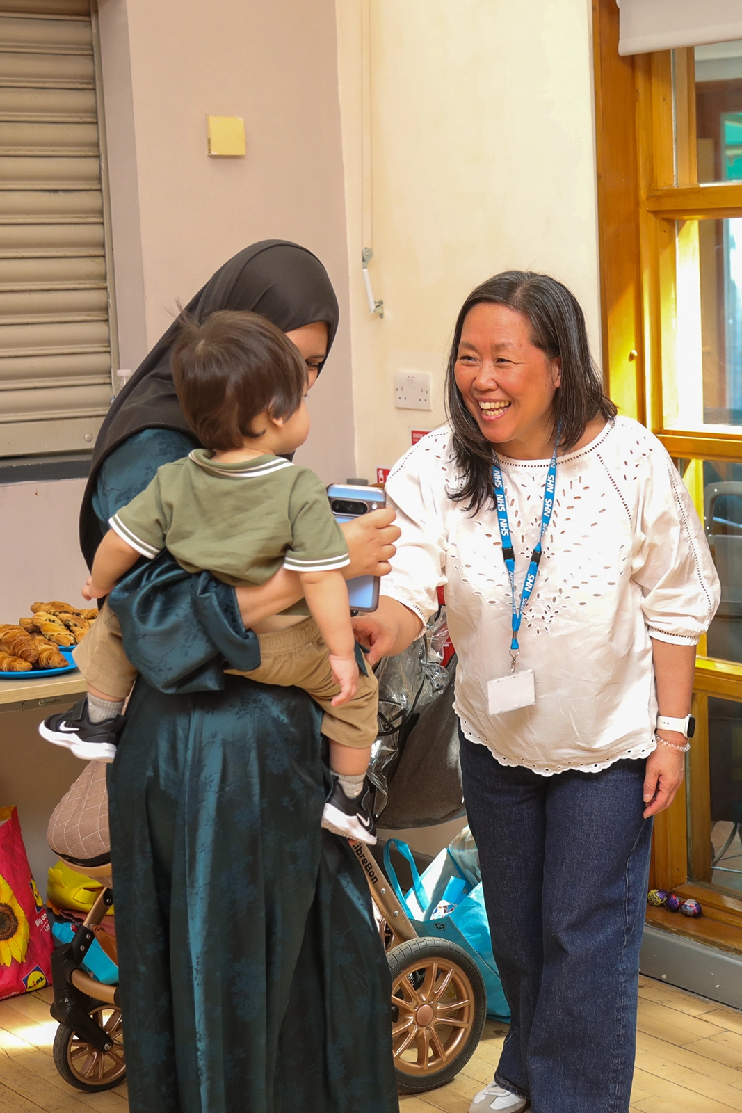 A woman wearing an NHS lanyard smiling and interacting with a woman holding a child dressed in green inside a room with a stroller and pastries on a table.