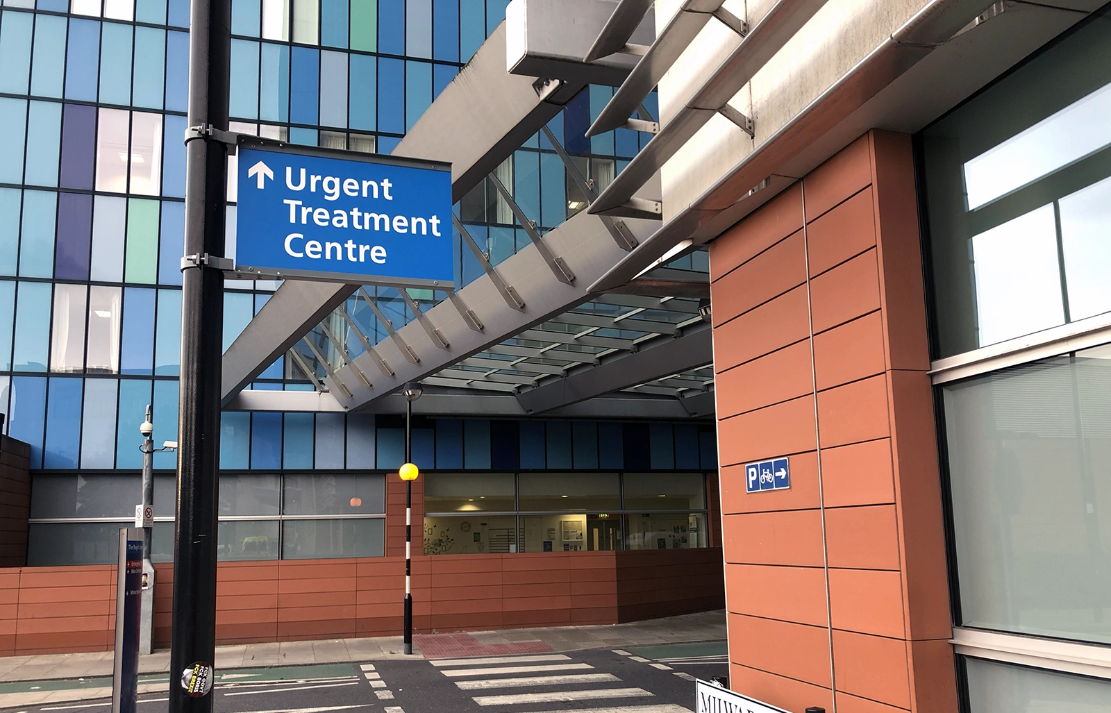 Blue directional sign pointing to the Urgent Treatment Centre outside a modern hospital building.