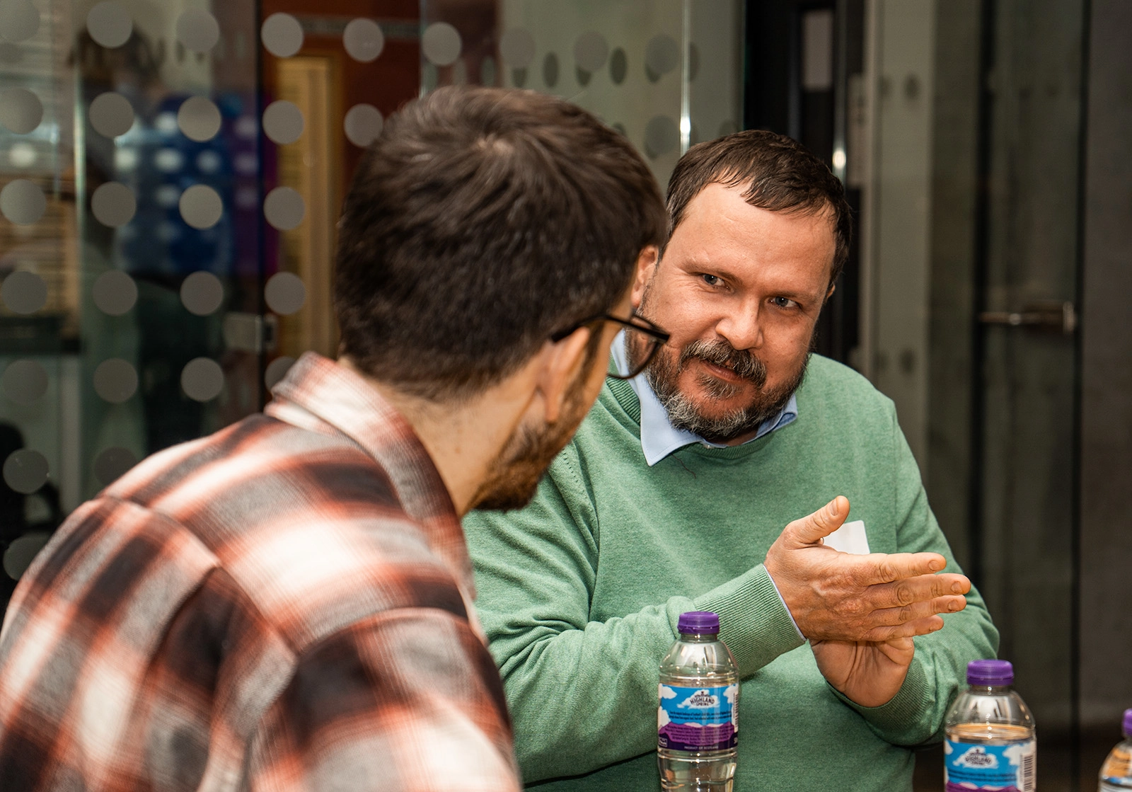 Two men engaged in a conversation at a table with bottled water, one wearing a green sweater and gesturing with hands.