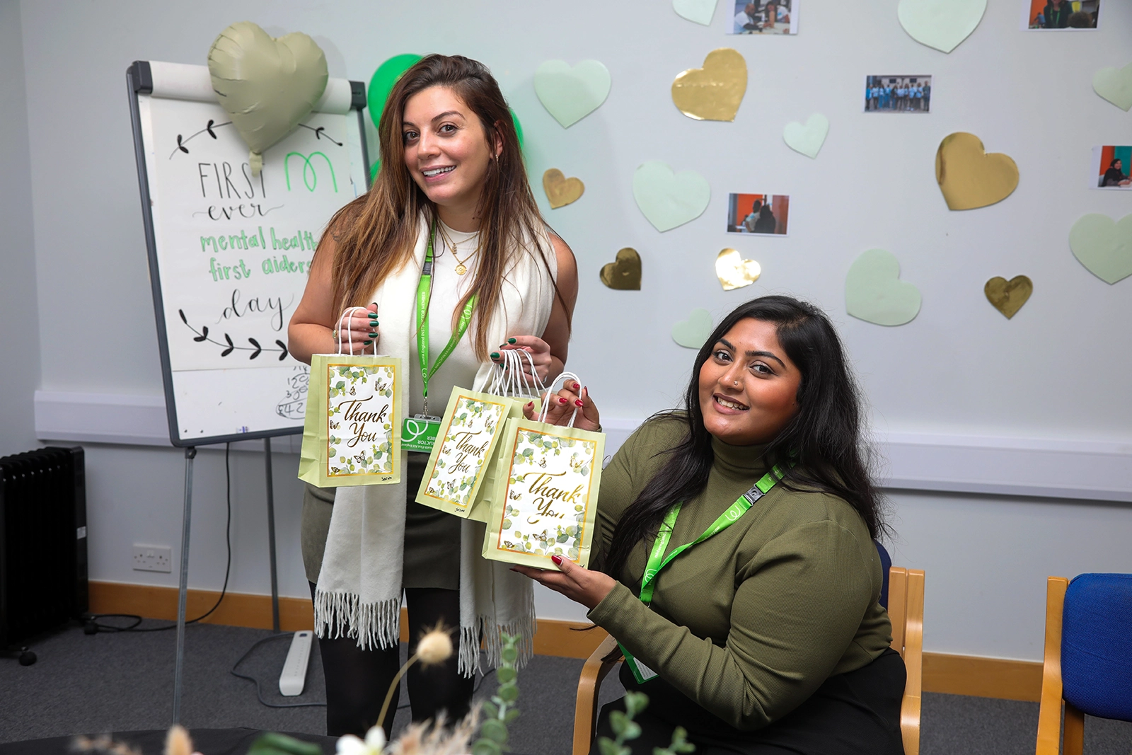 Two women smiling indoors holding thank you gift bags with heart decorations and a Board showing 'first ever mental health first aiders day' in the background.