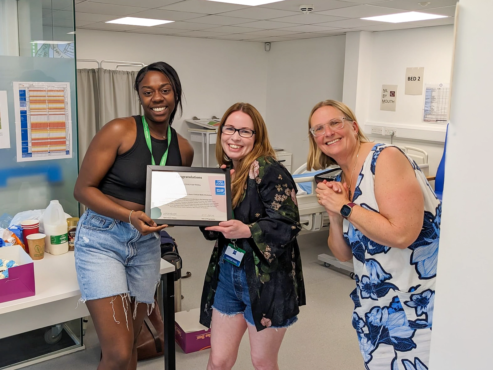 Three smiling women in a medical setting, one holding a framed certificate, with hospital beds and equipment in the background.