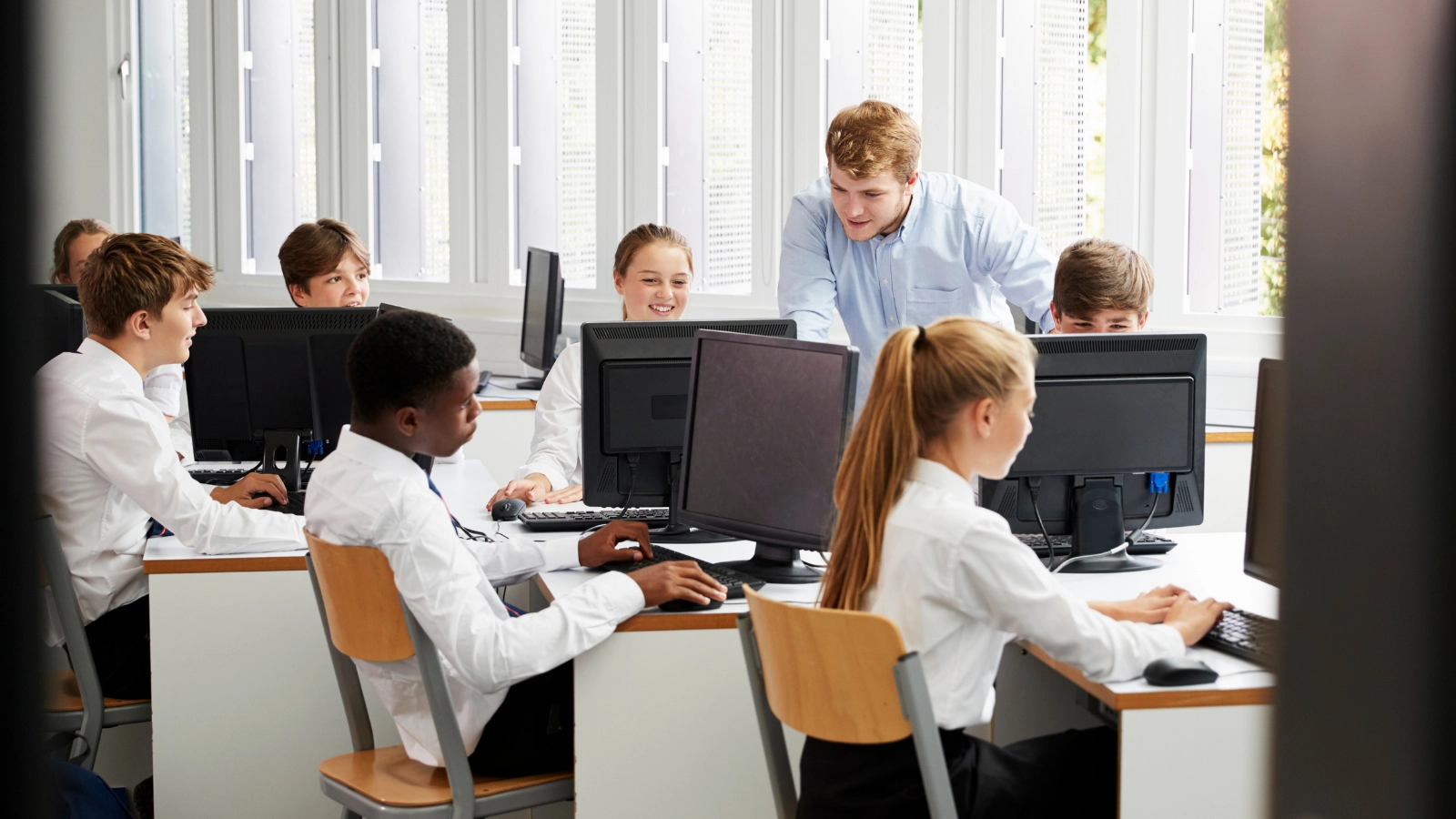 Teacher assisting a group of students working on desktop computers in a bright classroom.