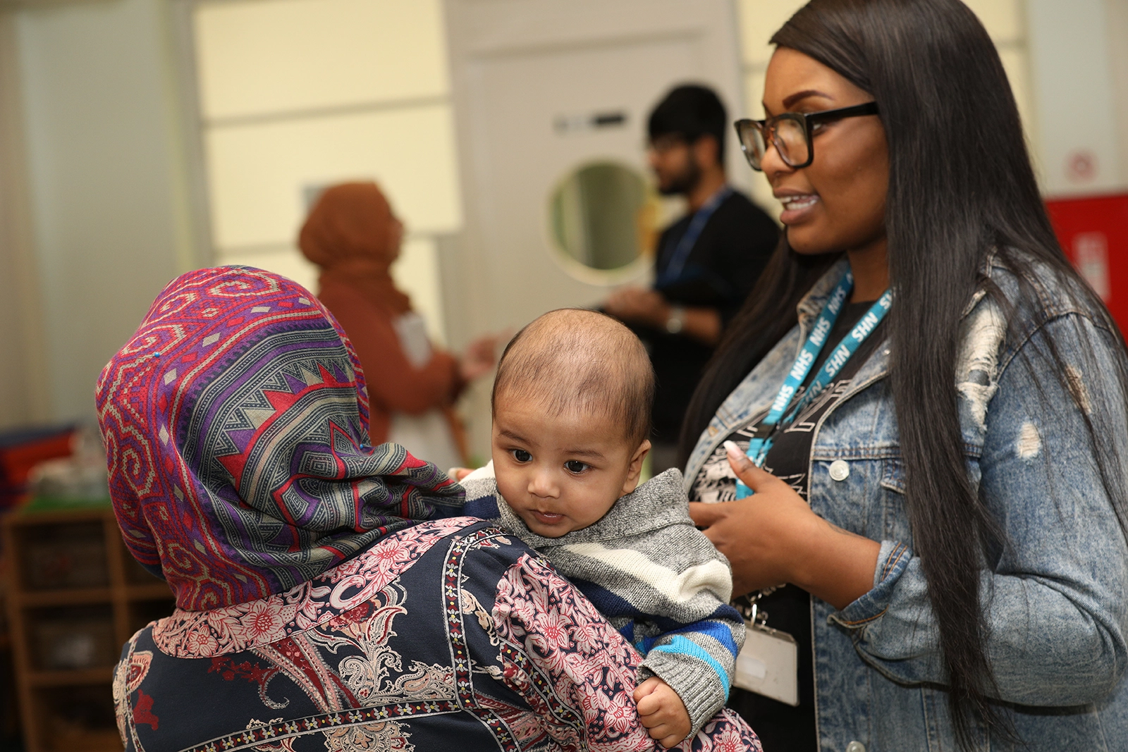 Woman in colorful headscarf holding a baby, talking to a smiling woman wearing glasses and a denim jacket with an NHS lanyard.