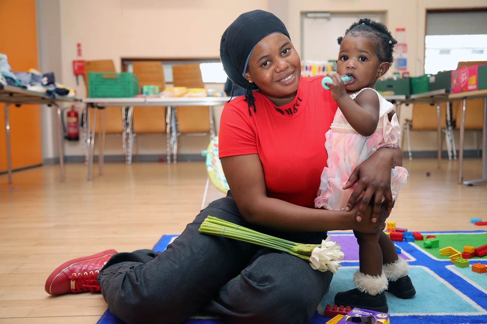Smiling woman in a red shirt sitting on the floor holding a baby girl in a pink dress who is chewing on a toy.