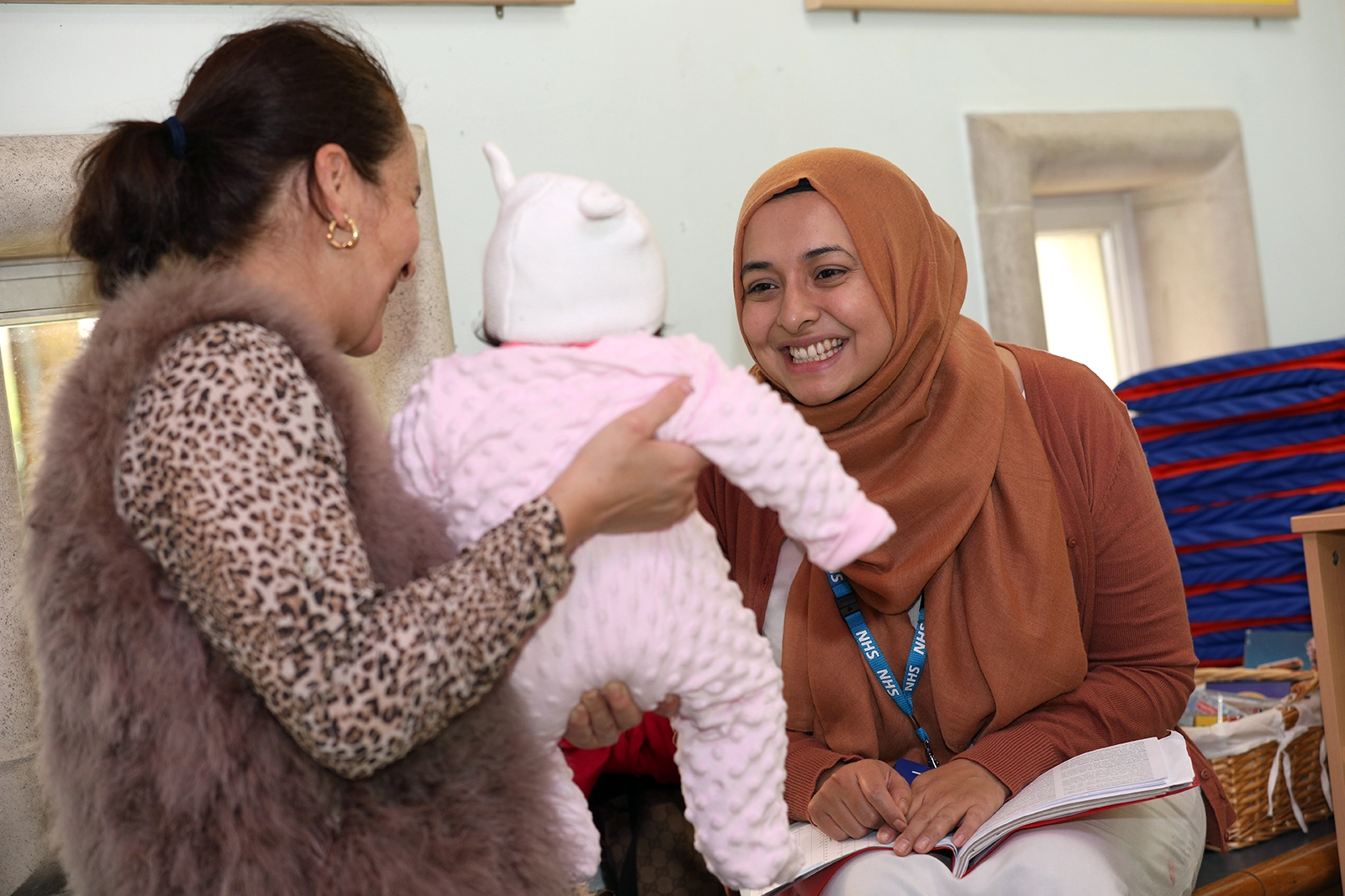 Smiling woman wearing a headscarf holding an open book interacts with a woman holding a baby dressed in pink.