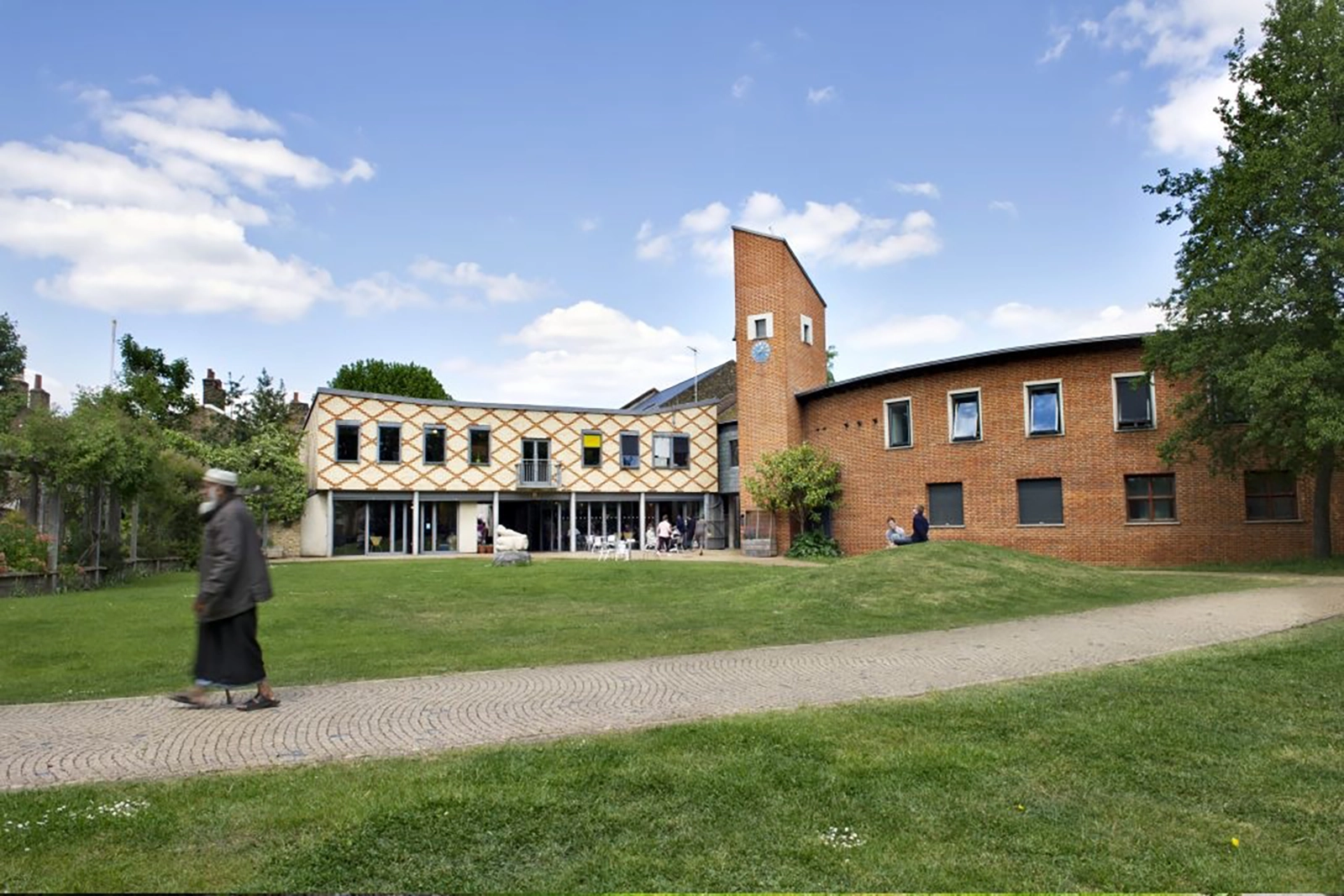 The Bromley by Bow center, a large community center building with brick and patterned exterior, a clock tower, people sitting outside, and a person walking on a path in front.