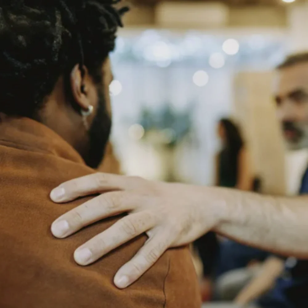 A person gently placing a hand on the shoulder of another person in a supportive gesture, with blurred background figures.