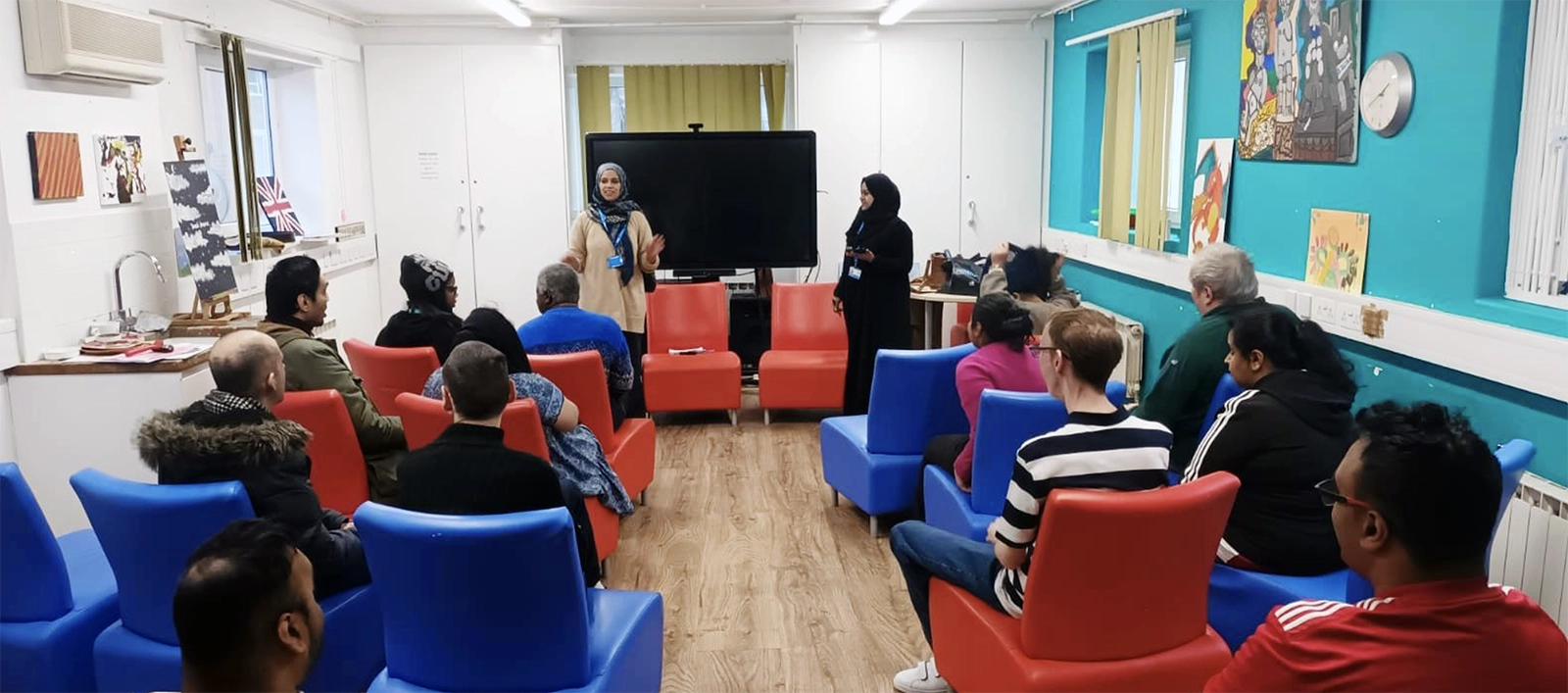 Diverse group of people seated on blue and red chairs in a room, listening to two women standing and speaking near a large screen.