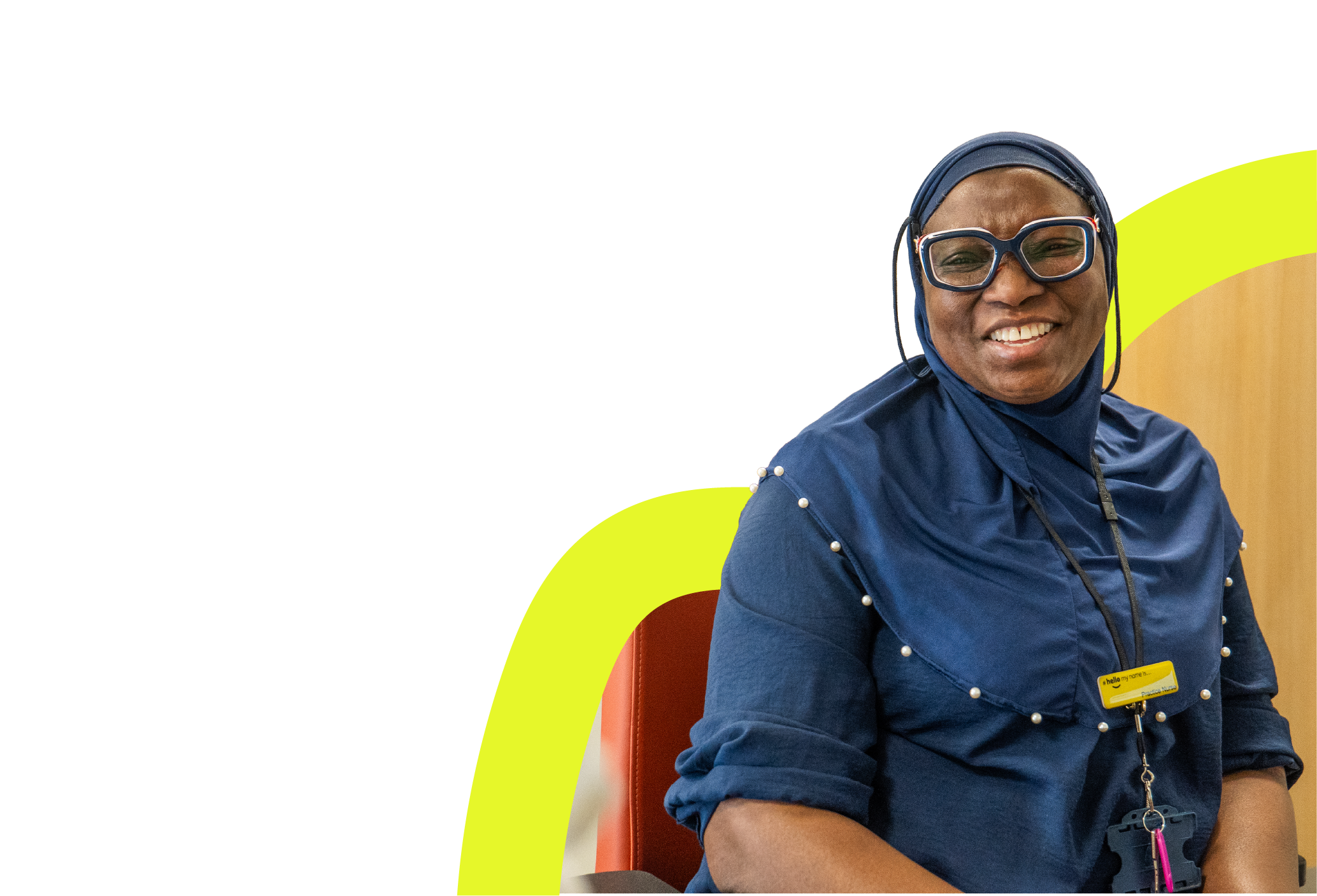 Smiling woman wearing glasses and a blue hijab with pearl details, sitting on a red chair.