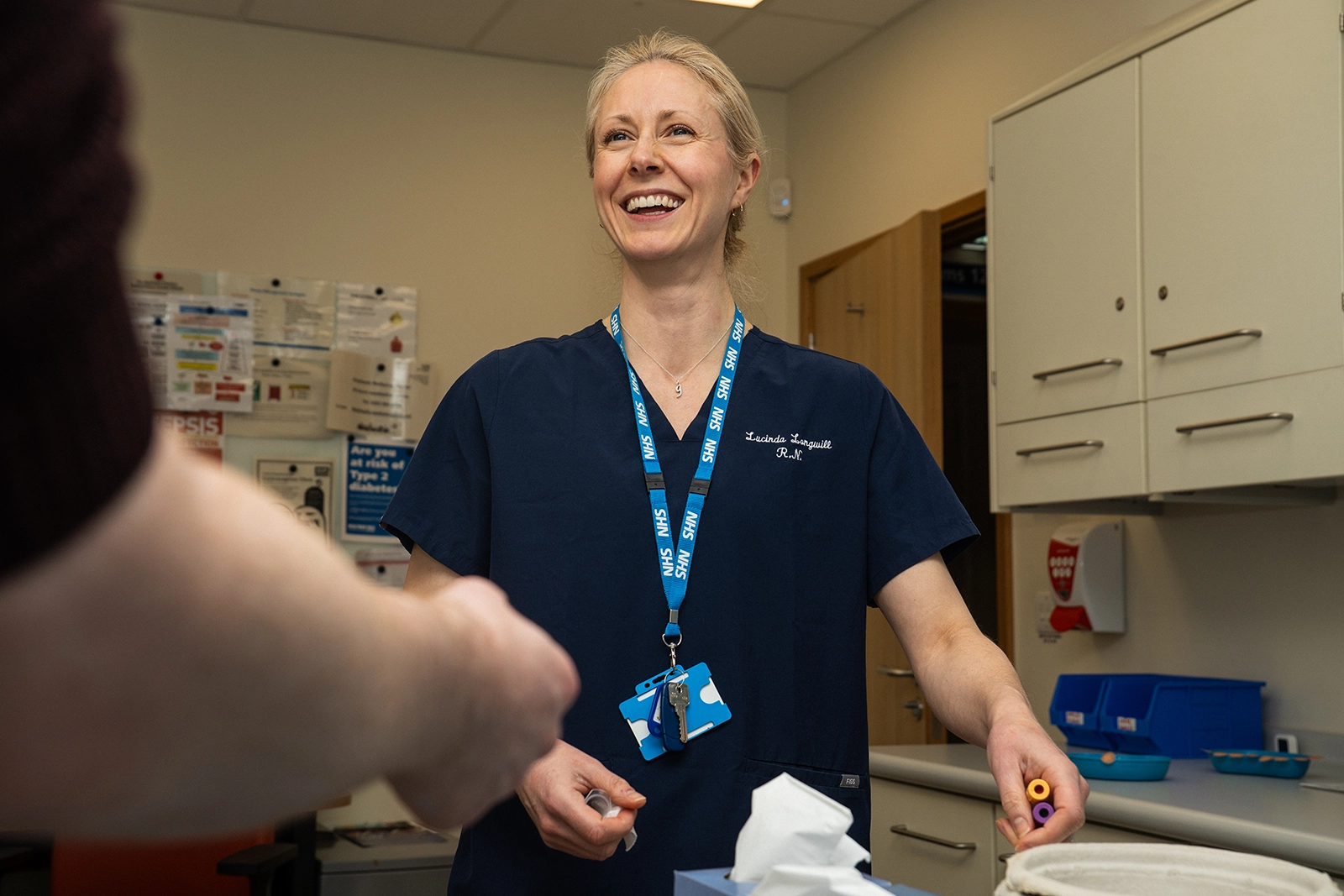 Smiling female nurse in navy scrubs with NHS lanyard handing medical supplies to another person.