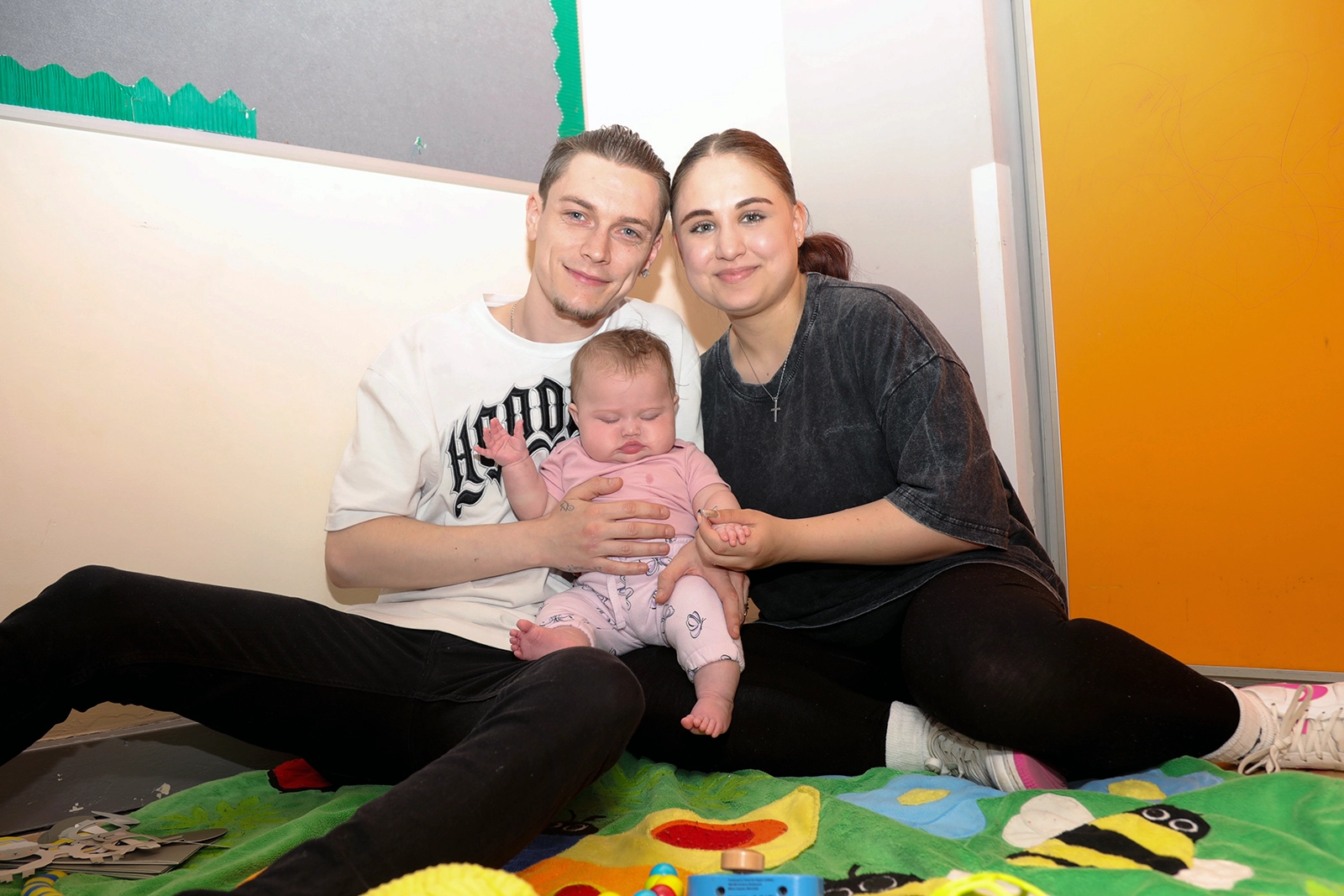 Young couple sitting on a colorful blanket holding a baby dressed in pink in a playroom.