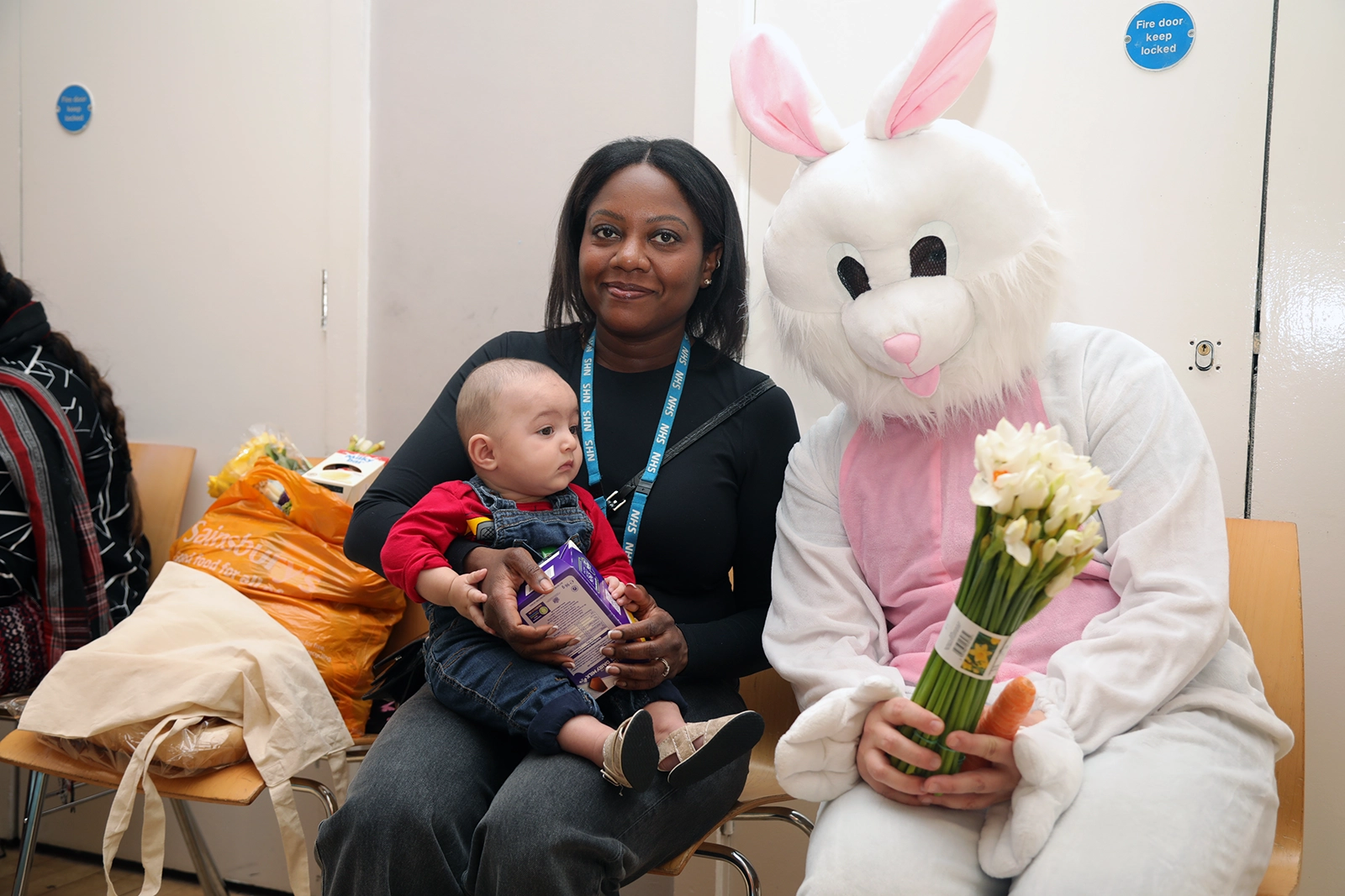A woman holding a baby sitting next to a person dressed in a white and pink Easter bunny costume holding a bouquet of white flowers and a carrot.