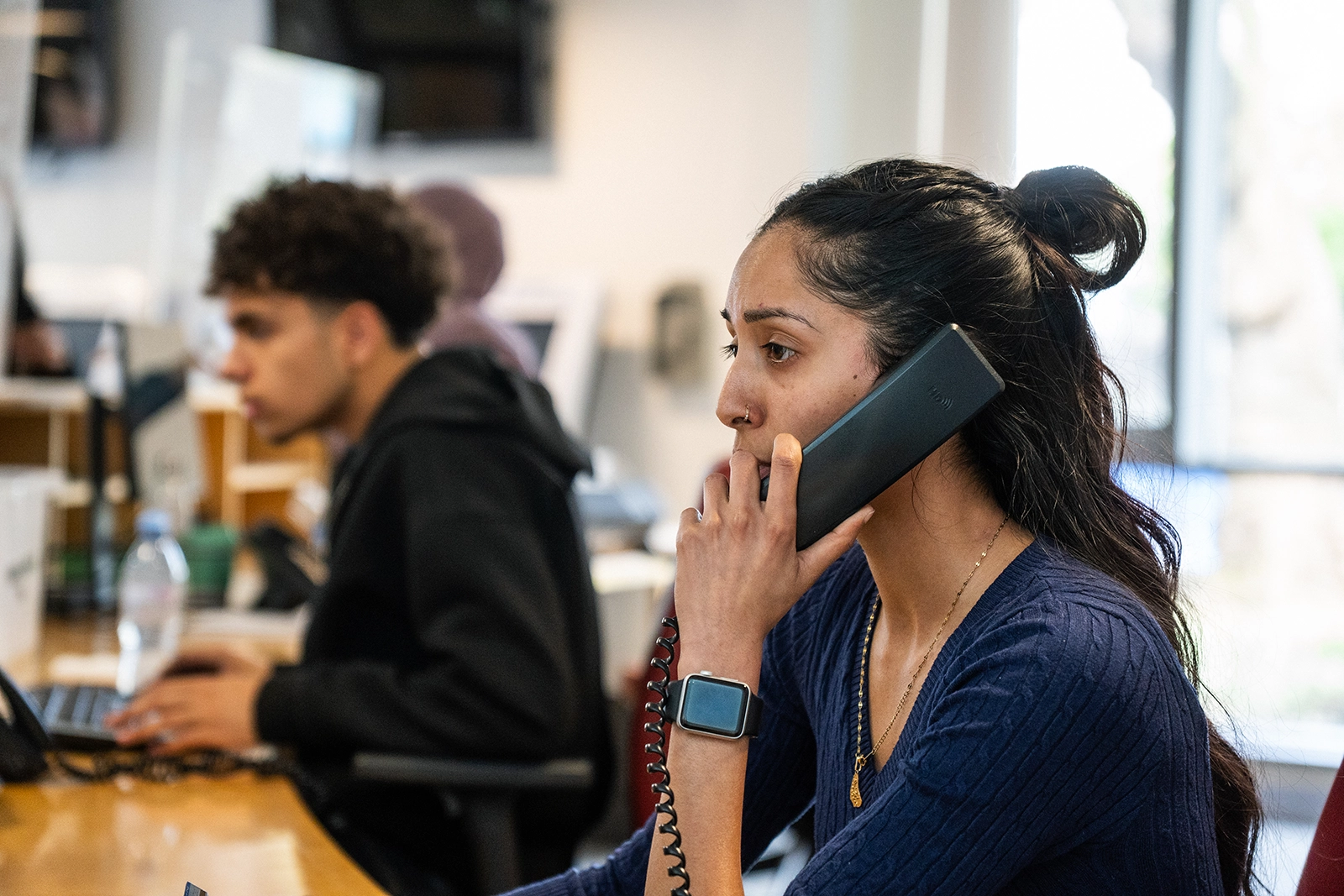 Young woman with a ponytail talking on a corded office phone while working at a desk in an office environment.