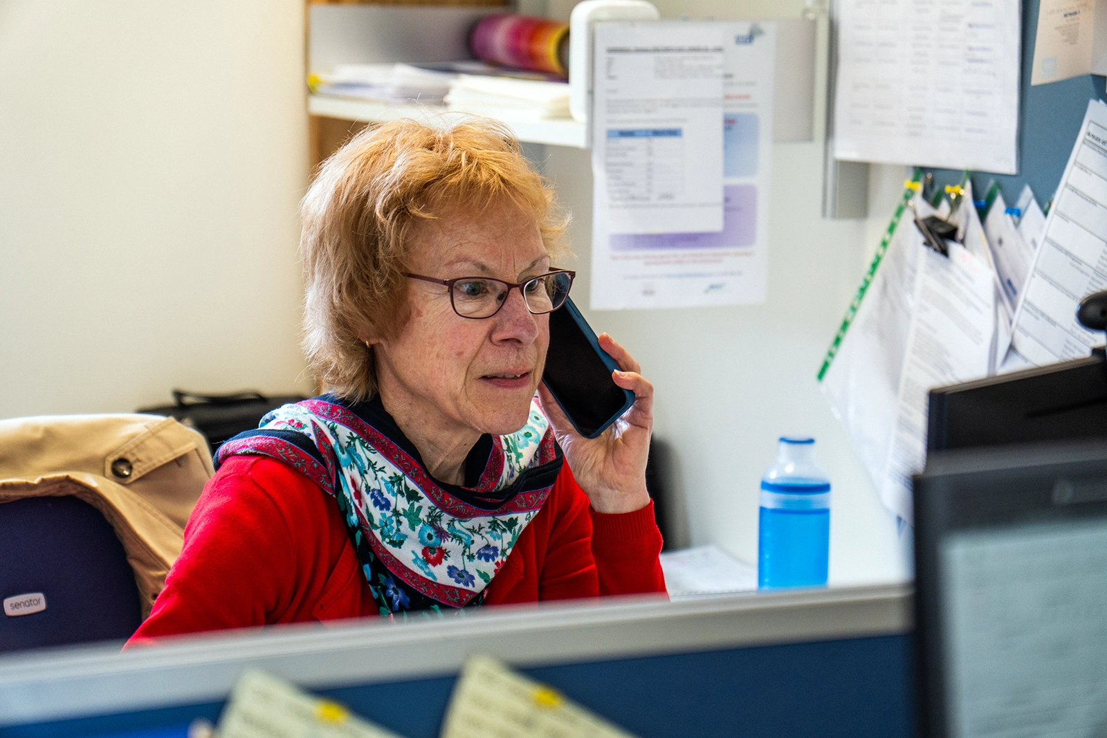 Middle-aged woman wearing glasses and a red sweater talking on a smartphone in an office.