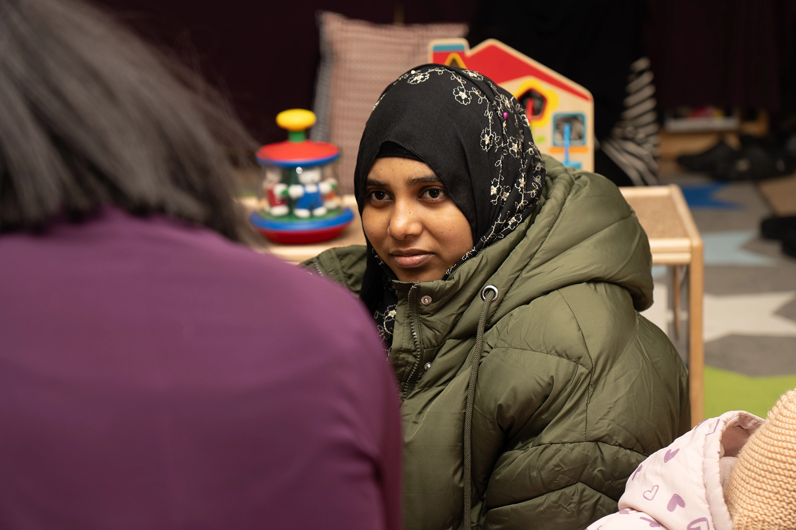 Woman wearing a black floral hijab and green puffer jacket looking attentively at another person in a purple top indoors.