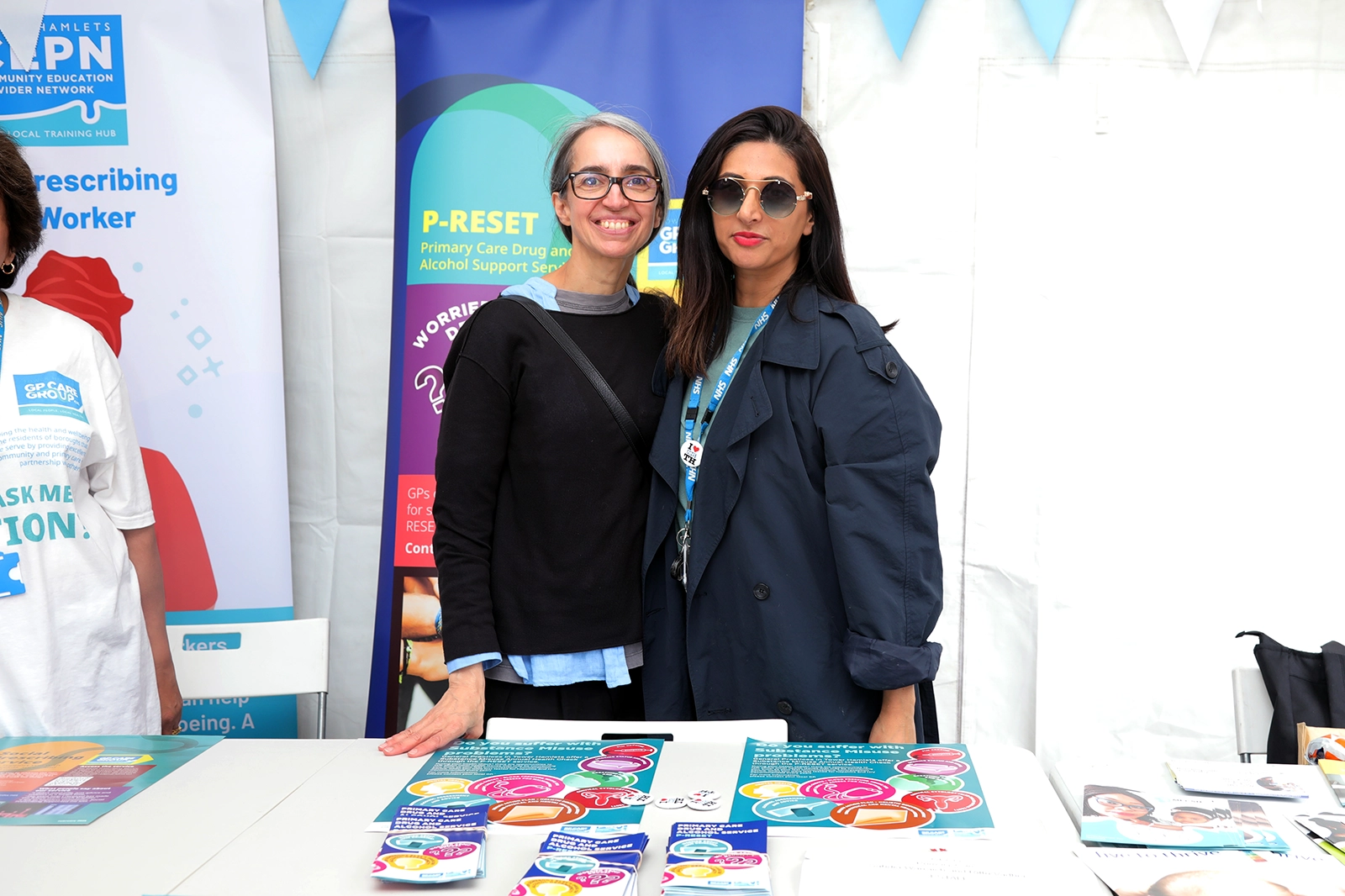 Two women standing behind a table with informational pamphlets, smiling at the camera; one woman wears glasses and a black sweater, the other wears sunglasses and a dark blue coat with NHS lanyard, with colorful banners behind them related to primary care and support services.