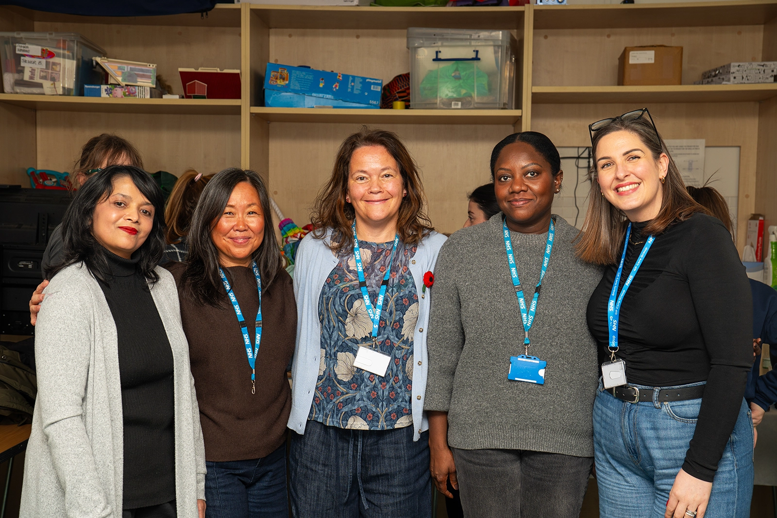 Five women standing indoors in front of shelves filled with storage boxes and items, all wearing NHS lanyards and smiling at the camera.